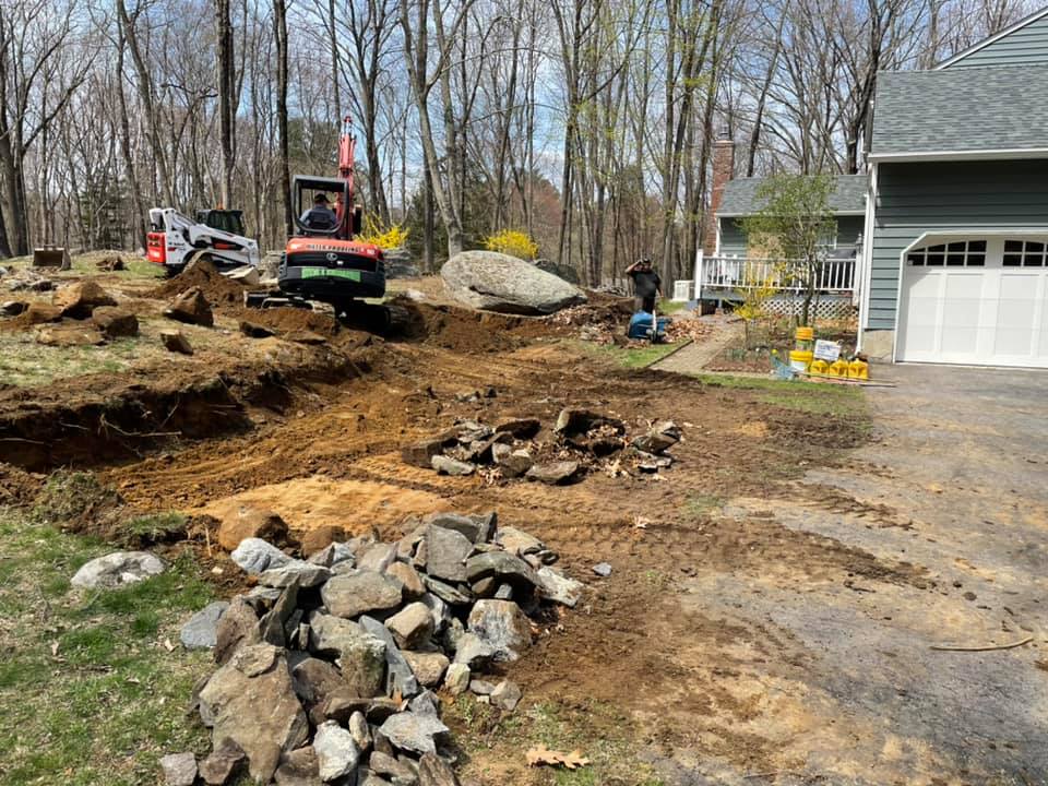 Construction site with excavators, rocks, and dirt near a house with a garage.