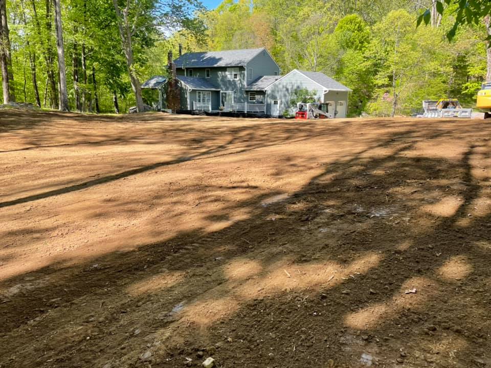 Cleared land in front of a gray house, ready for landscaping; trees and blue sky visible.