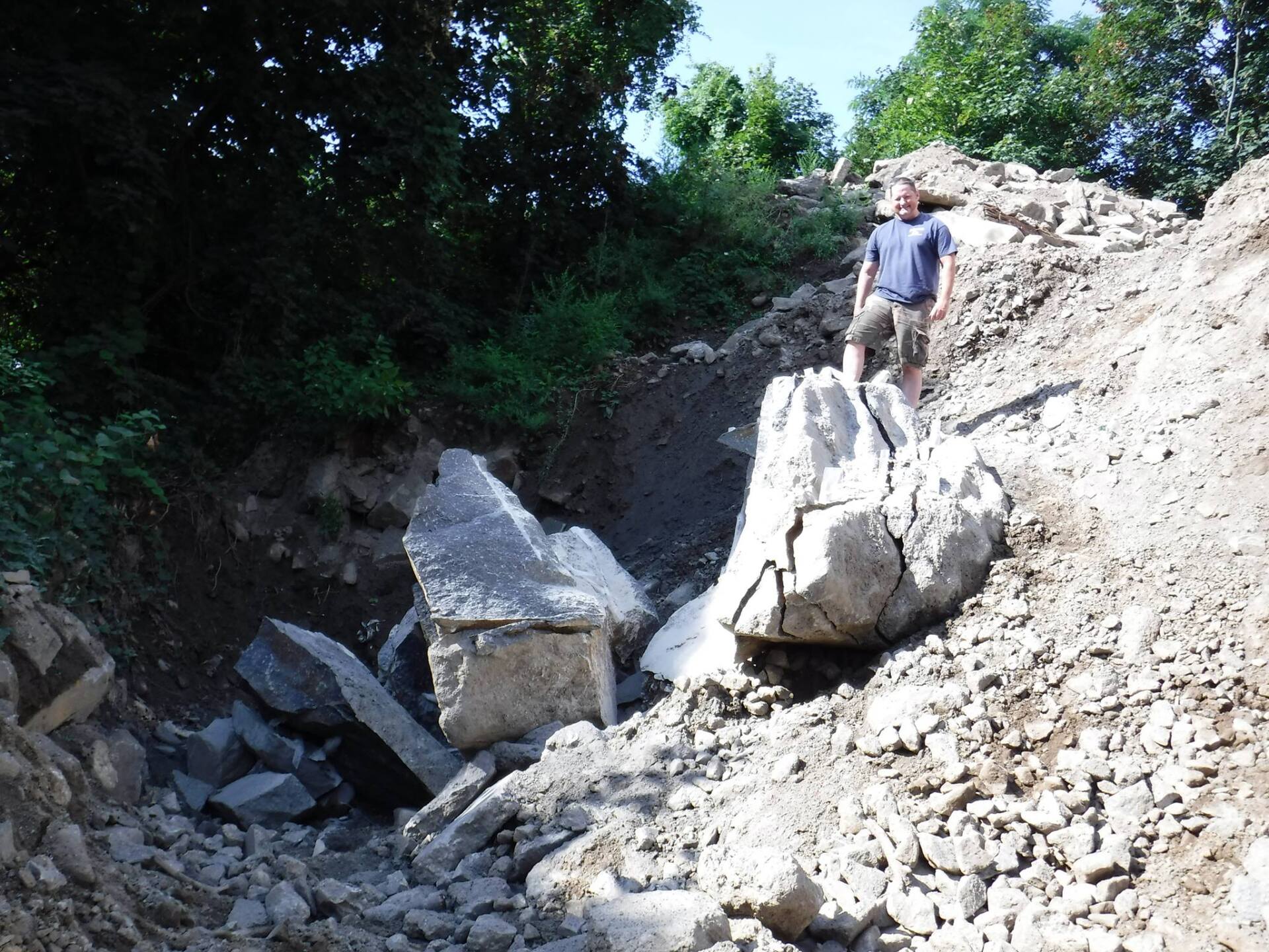 Man standing near large broken concrete blocks in a dirt ditch. Green trees in background.