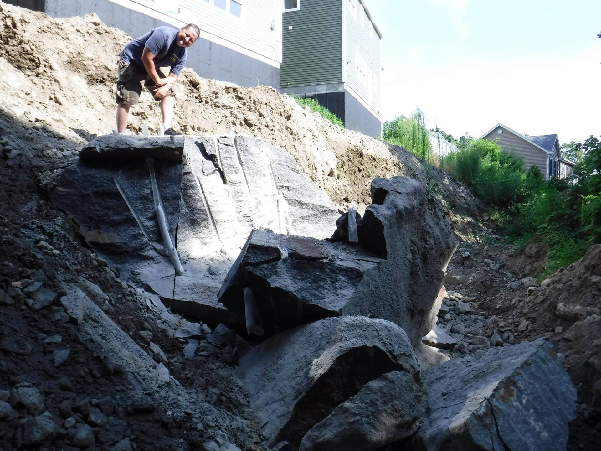 Man excavating large rocks from a deep trench. Dirt surrounds the rocks; building in background.
