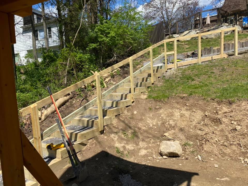 Wooden staircase with handrails on a hillside, leading up to a fence. Sunny day.
