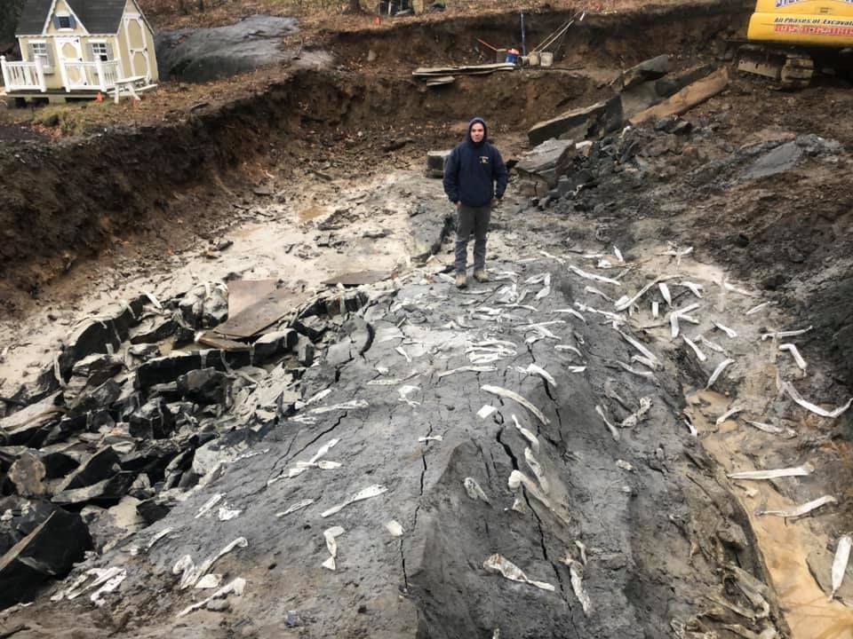 Man stands in large excavation pit filled with rock and white fragments. Small house in background.