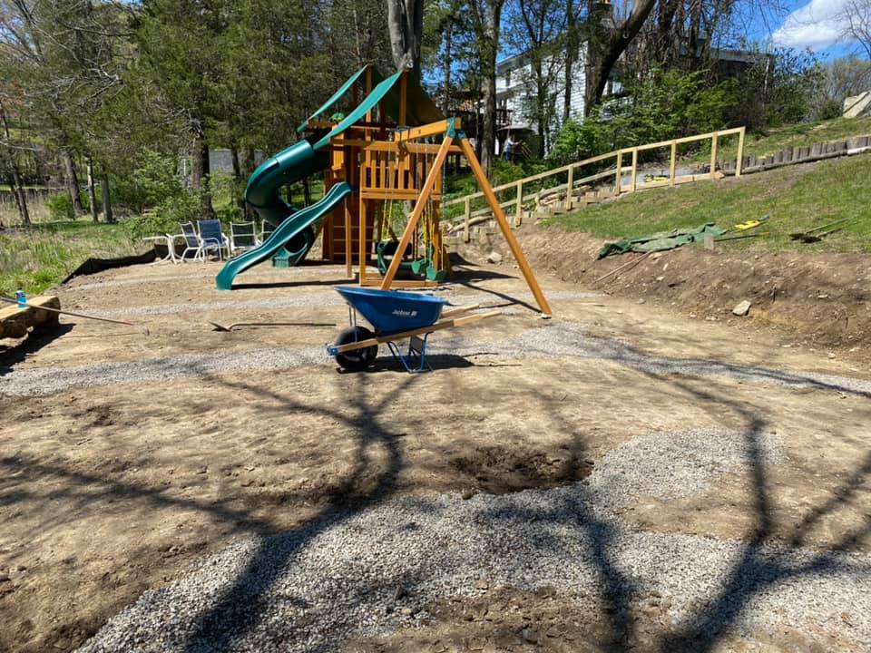 Playground with slide and swings in a yard under construction; wheelbarrow, gravel, and hillside in the background.