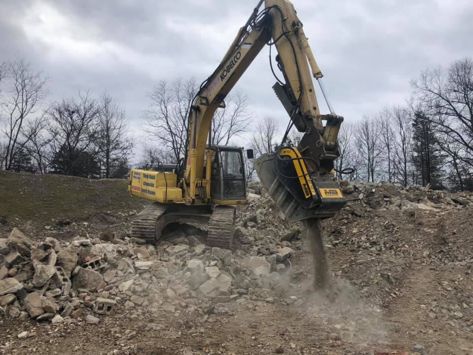 Yellow excavator with a demolition head breaking up concrete debris in a construction zone.