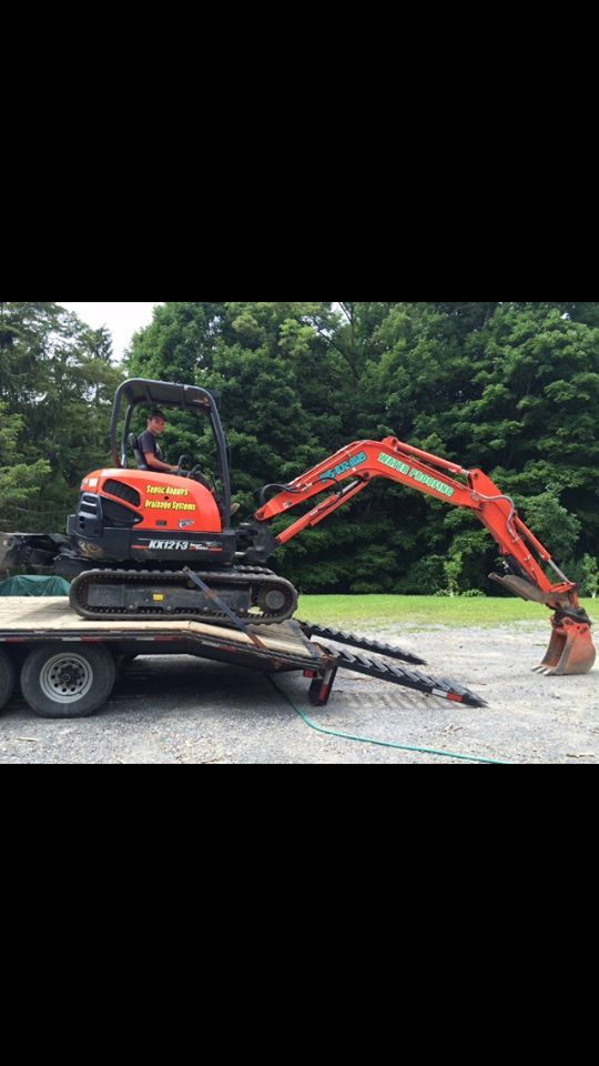 Orange Kubota mini-excavator on a trailer, with augers in front, set against a backdrop of trees.