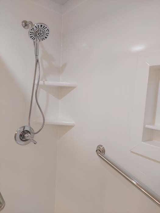 White accessible shower with handheld showerhead, corner shelves, and a grab bar.