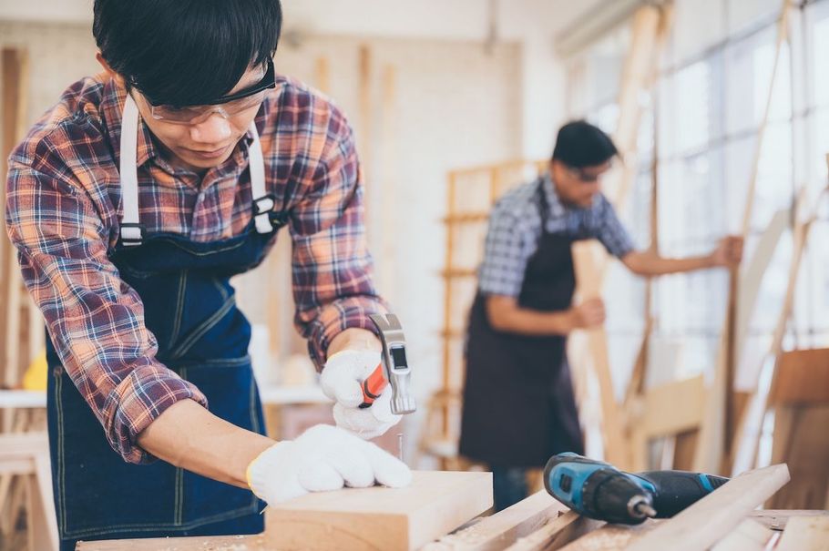 Two people woodworking in a bright workshop, one sanding a wooden board in the foreground.
