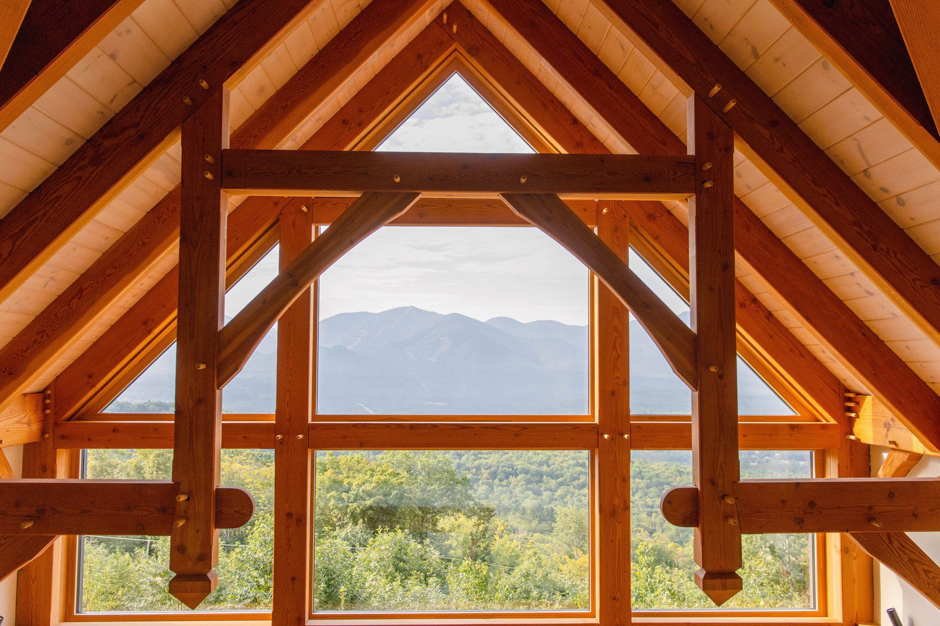 Wooden A-frame window framing a mountain landscape with trees and blue sky