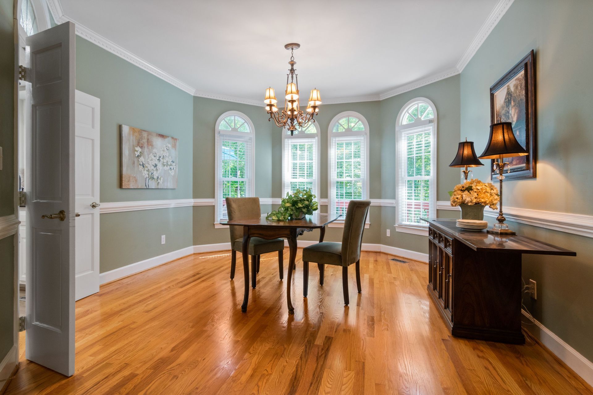 Dining room with hardwood floors, light blue walls, chandelier, and table by arched windows.