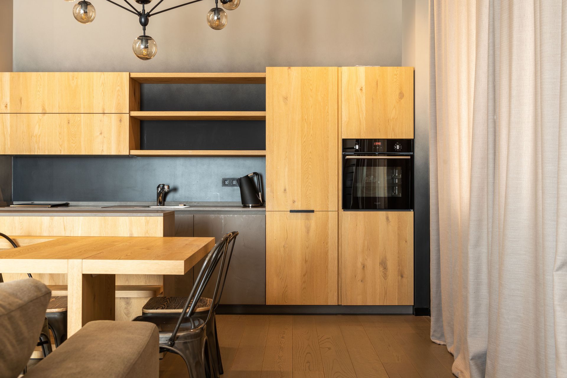 Modern kitchen with light wood cabinets, built-in oven, dining table, and sheer curtain by a window