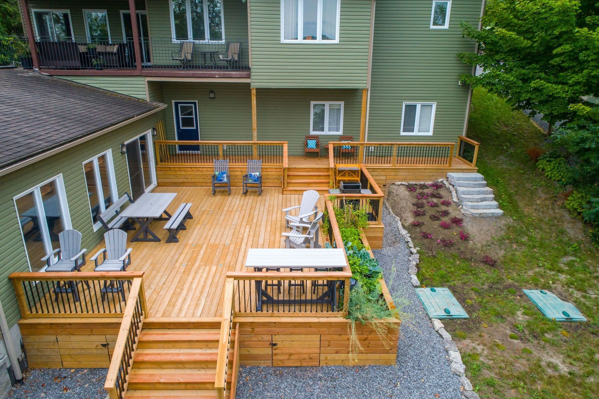 Wooden backyard deck with stairs, benches, and seating beside a green house, viewed from above