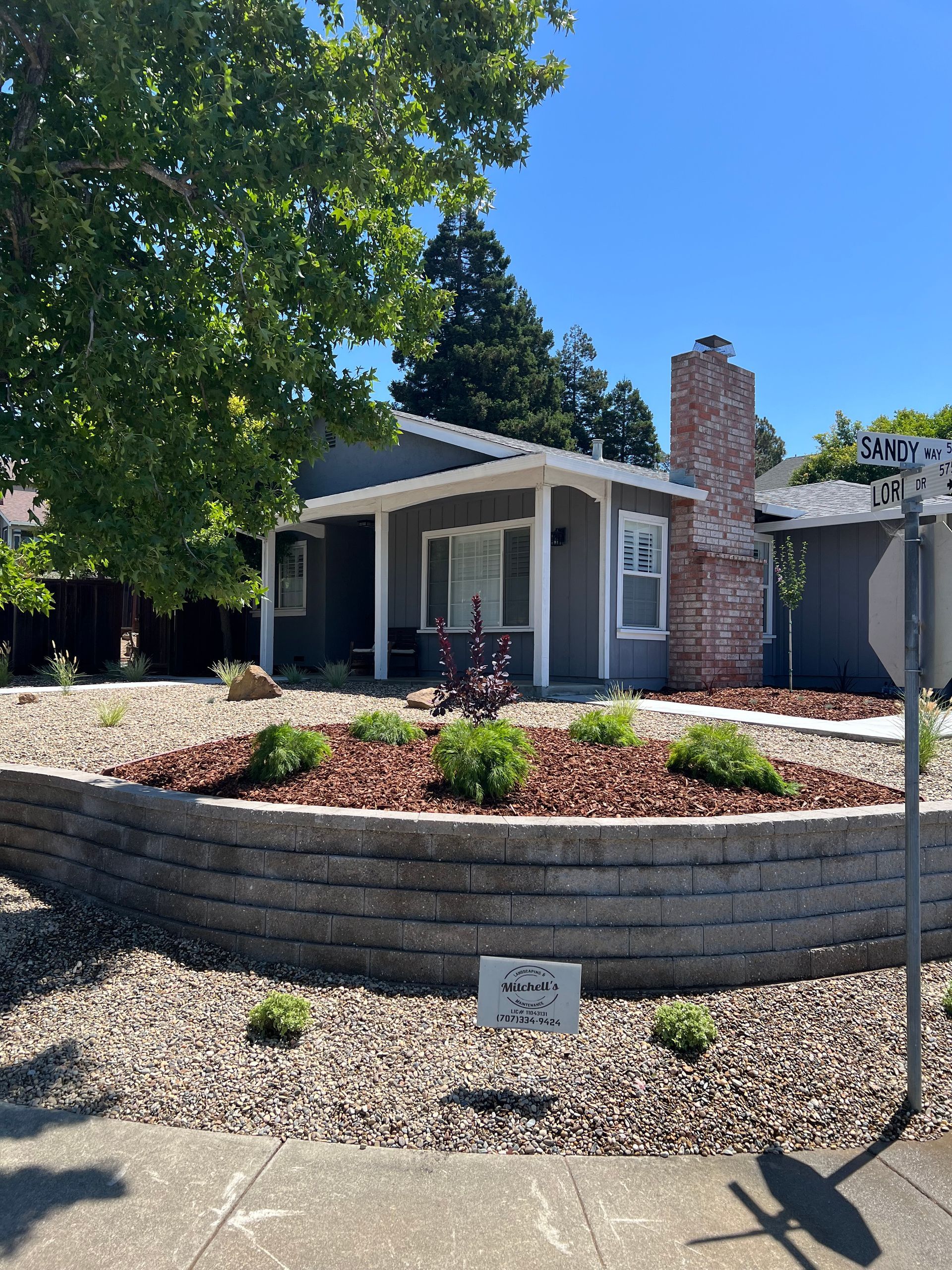 Gray house with a brick chimney, raised garden bed, and a sign on a sunny street.