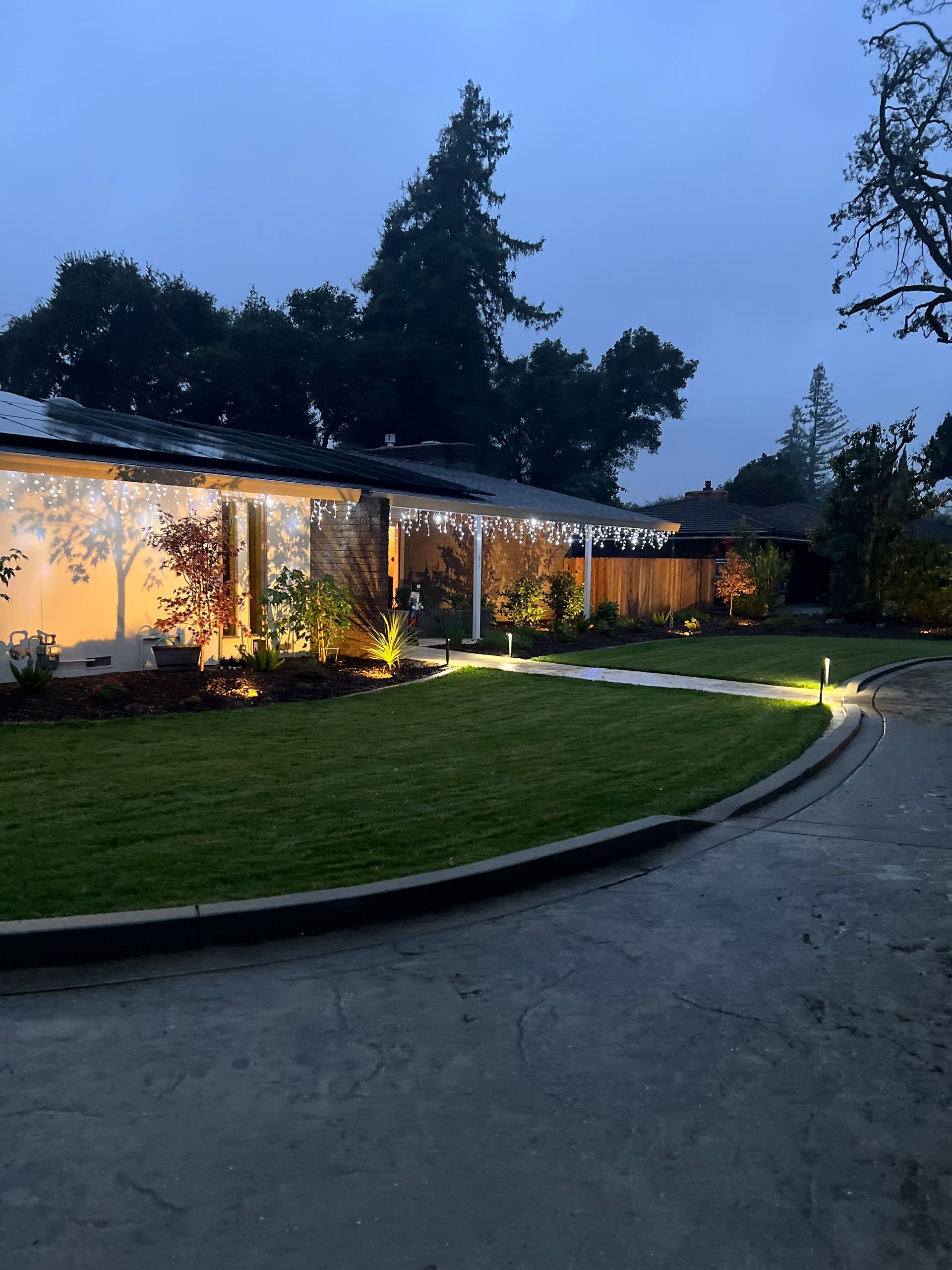 A house at dusk with string lights, lit landscaping, and a curved driveway.
