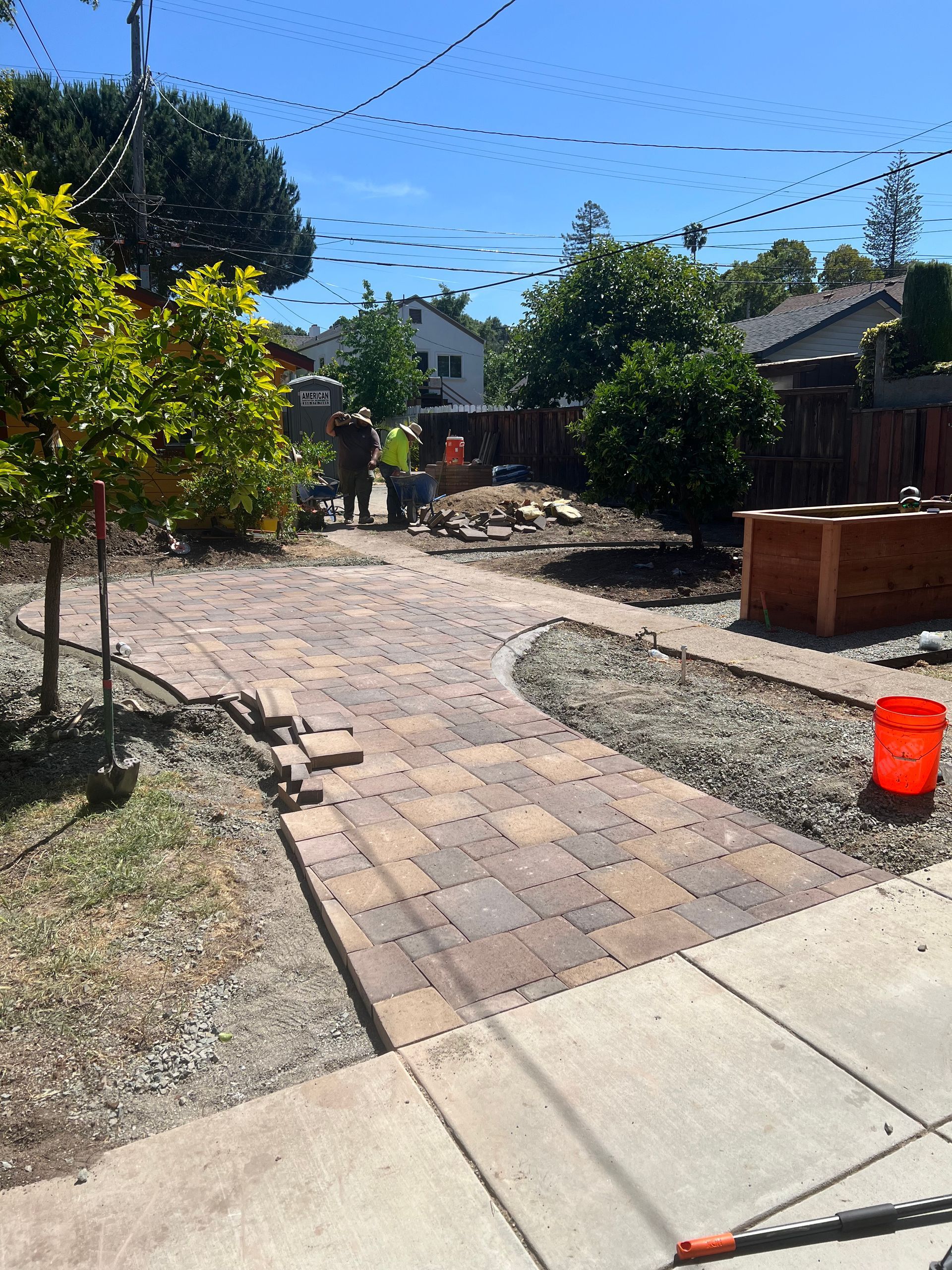Brick pathway under construction in a yard, with workers, trees, and an orange bucket.