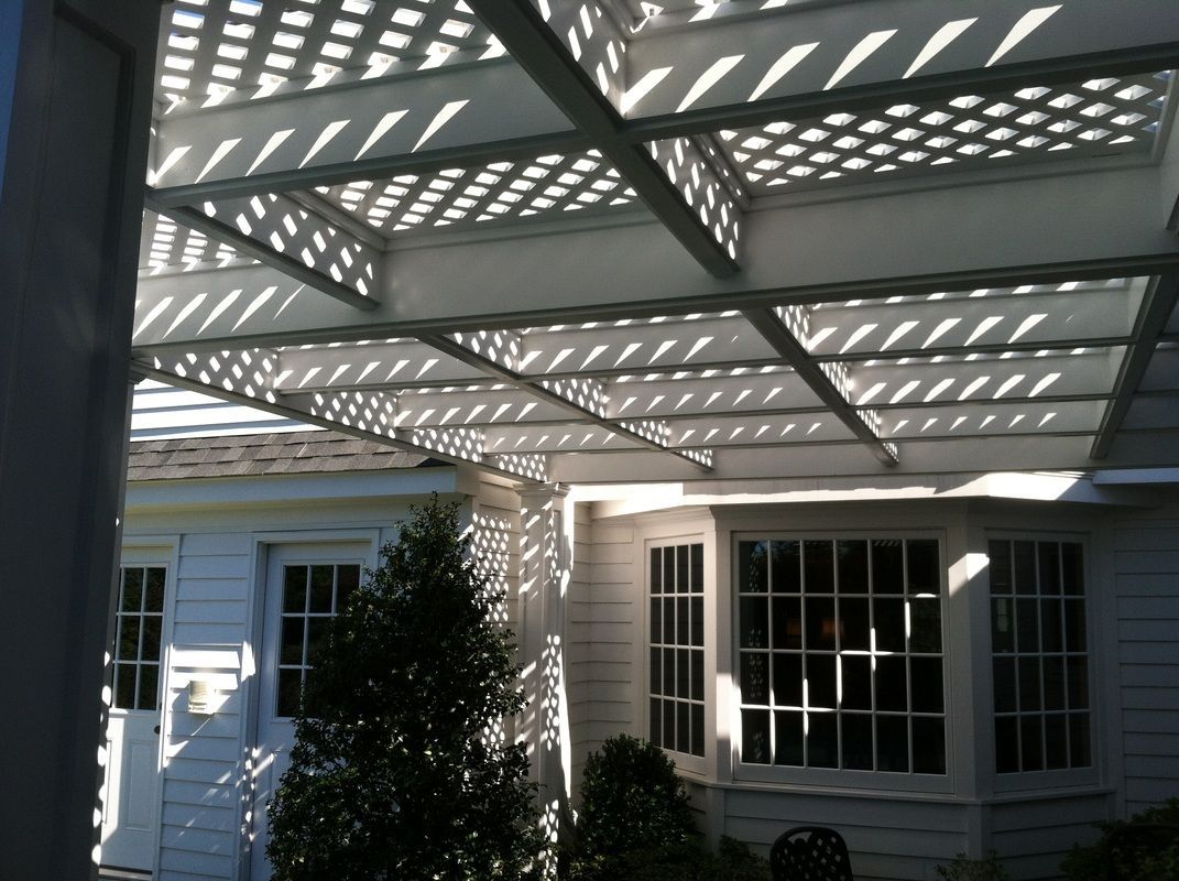 White pergola casting shadow patterns over a white house with windows and a small tree.