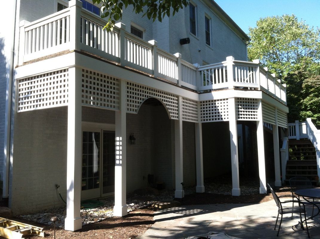 Two-story white deck with latticework and railing, supported by columns over a patio.