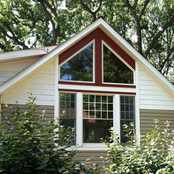 Gabled house with tan siding, burgundy trim around large windows, surrounded by green trees.