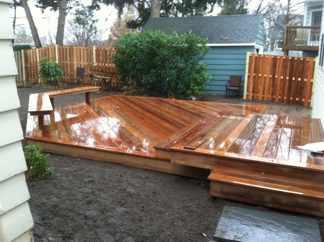 Wooden deck with chevron pattern, steps, and built-in bench in a backyard, surrounded by a fence and a small building.