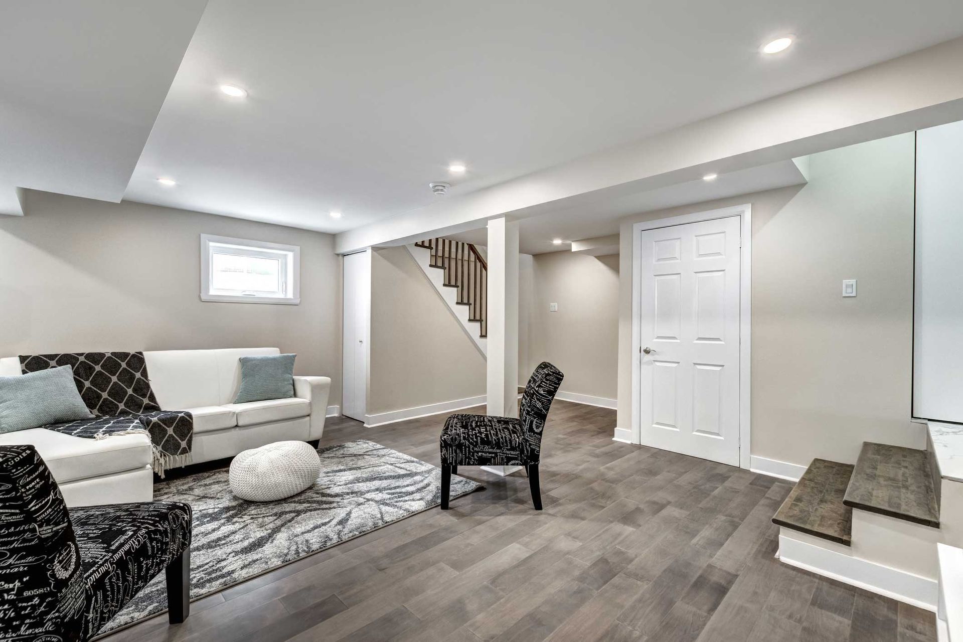 Finished basement living space with sofa, chairs, rug, stairs, and door. Gray wood floors, white walls, and recessed lighting.