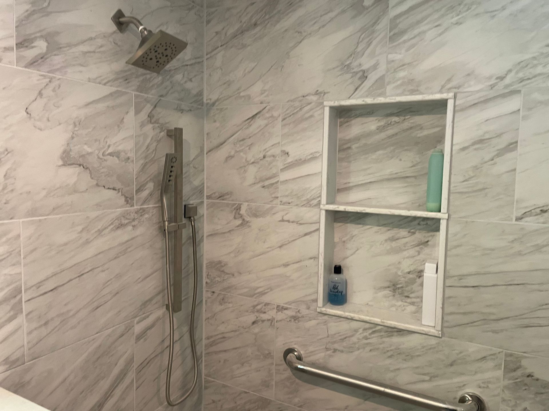Shower with gray marble-look tile, showerhead, handheld sprayer, built-in shelves with products, and grab bar.