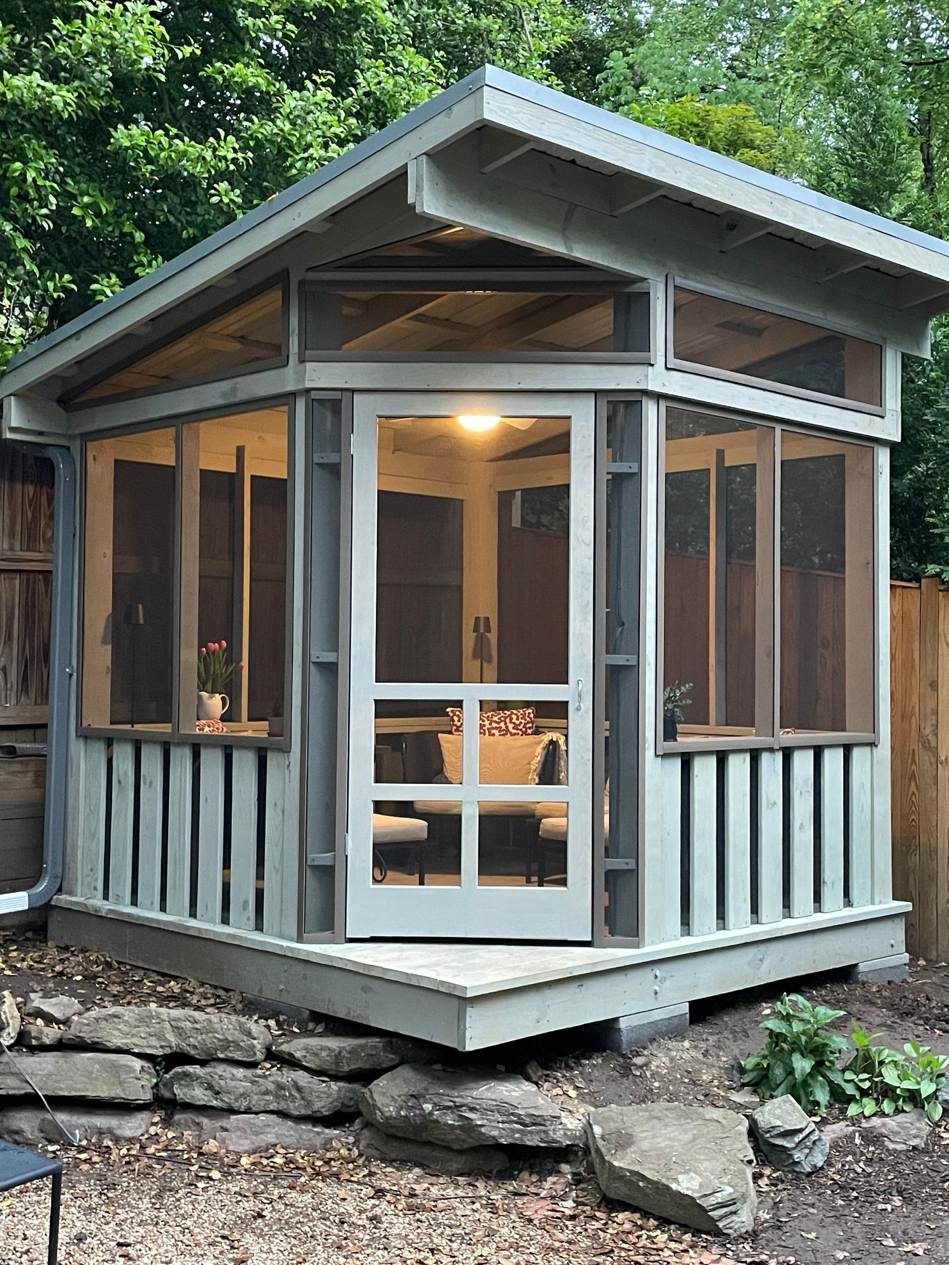 Screened-in gazebo with a light gray exterior, featuring a door, windows, and a raised wooden deck surrounded by rocks.