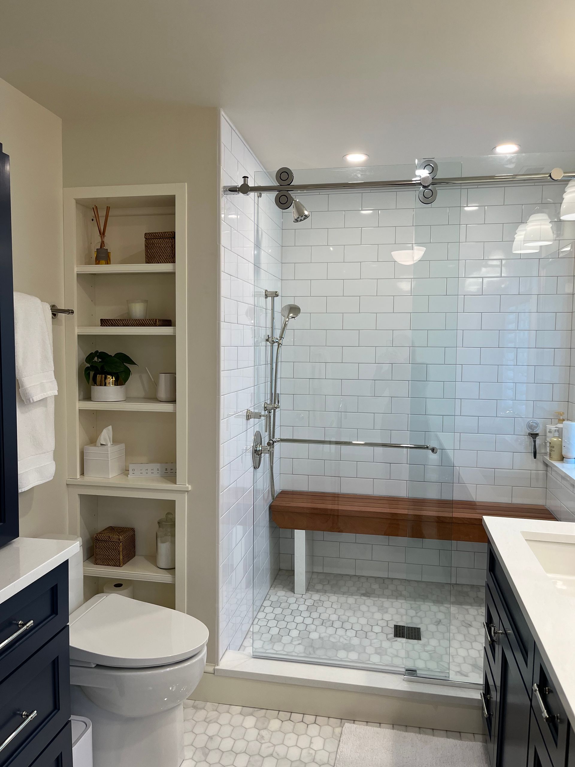 Bathroom with glass shower, white subway tile, wooden bench, built-in shelving, and navy vanity.