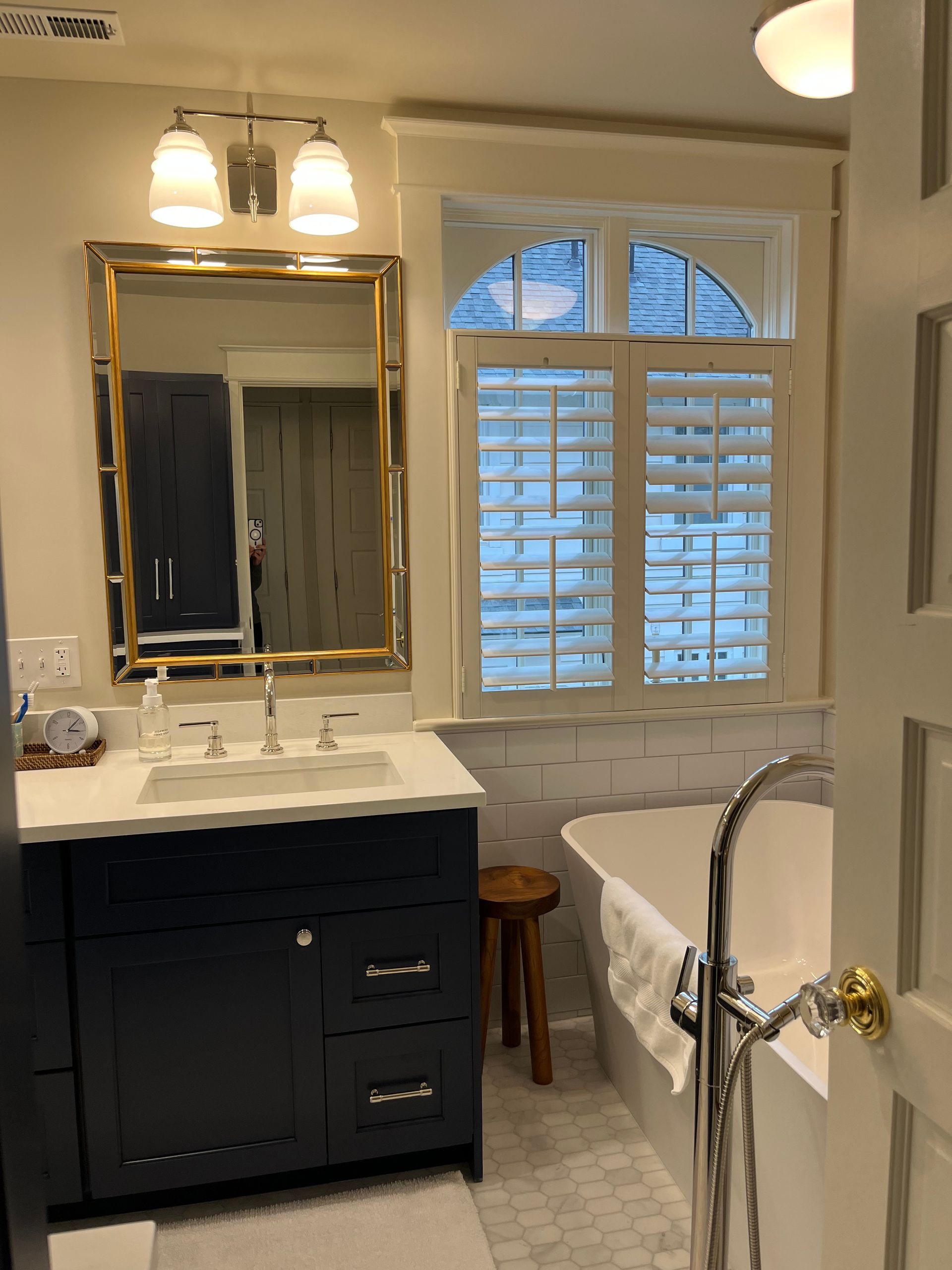Bathroom with navy vanity, white countertop, gold-framed mirror, window with shutters, and soaking tub.