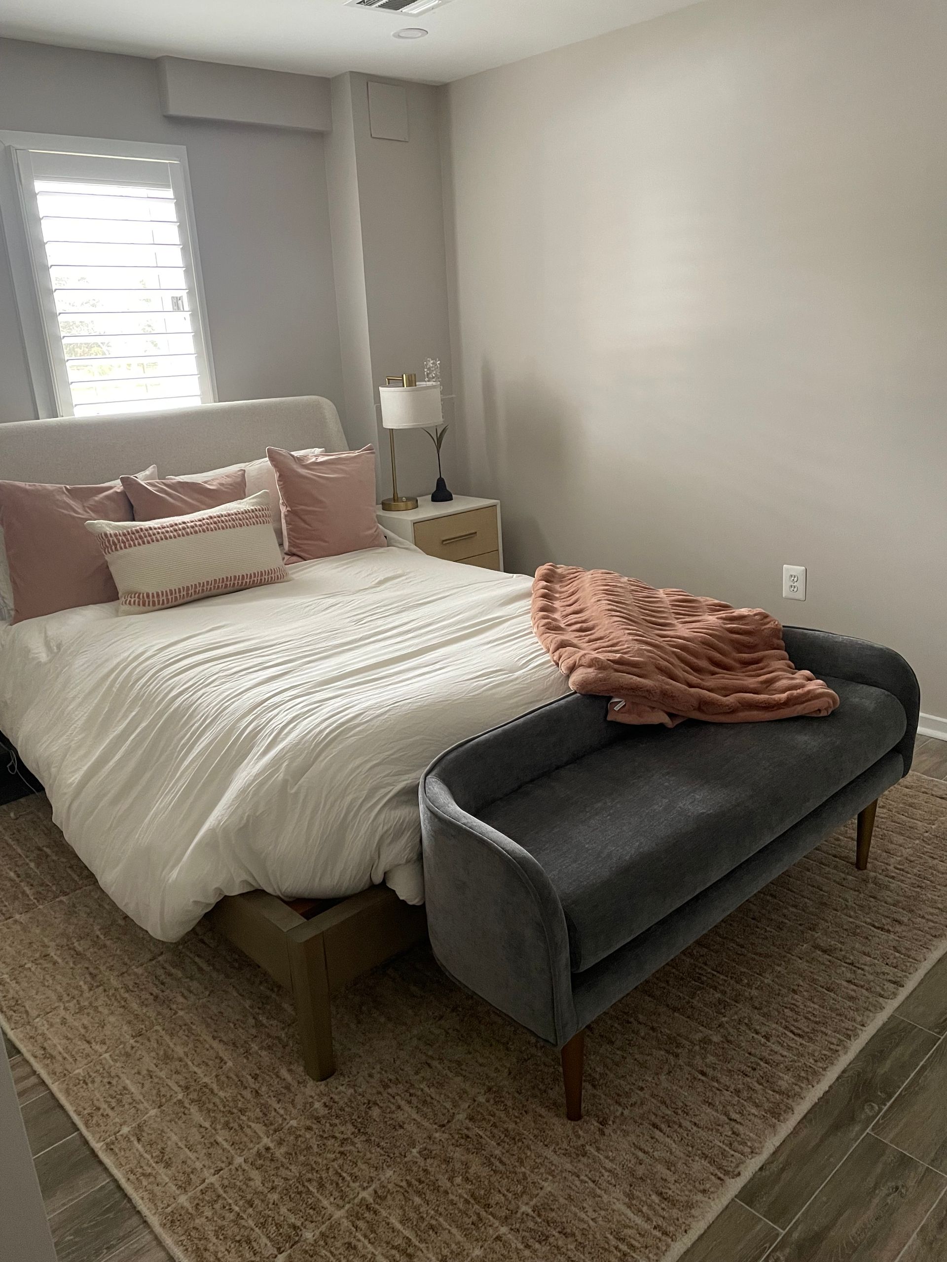 Bedroom with bed, bench, rug, and nightstand. Bed has white bedding and pink pillows. Grey bench at foot.