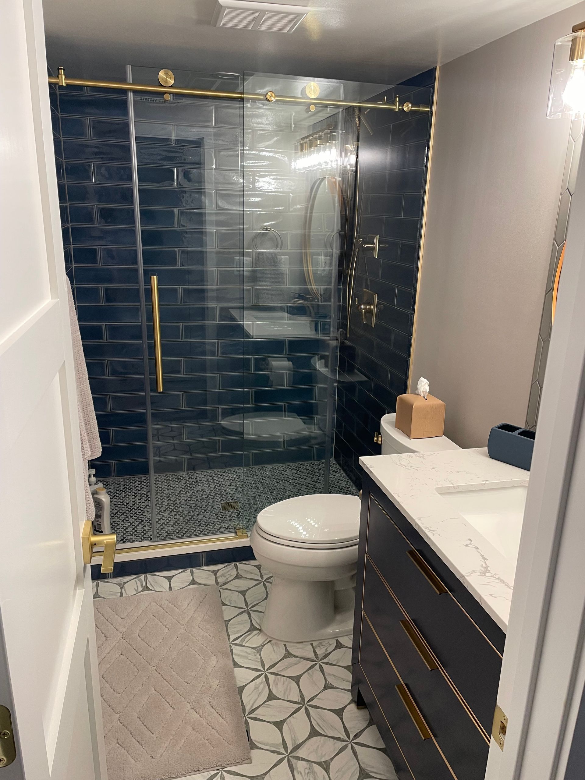 Bathroom with dark blue tiled shower, gold hardware, patterned floor, and dark vanity.