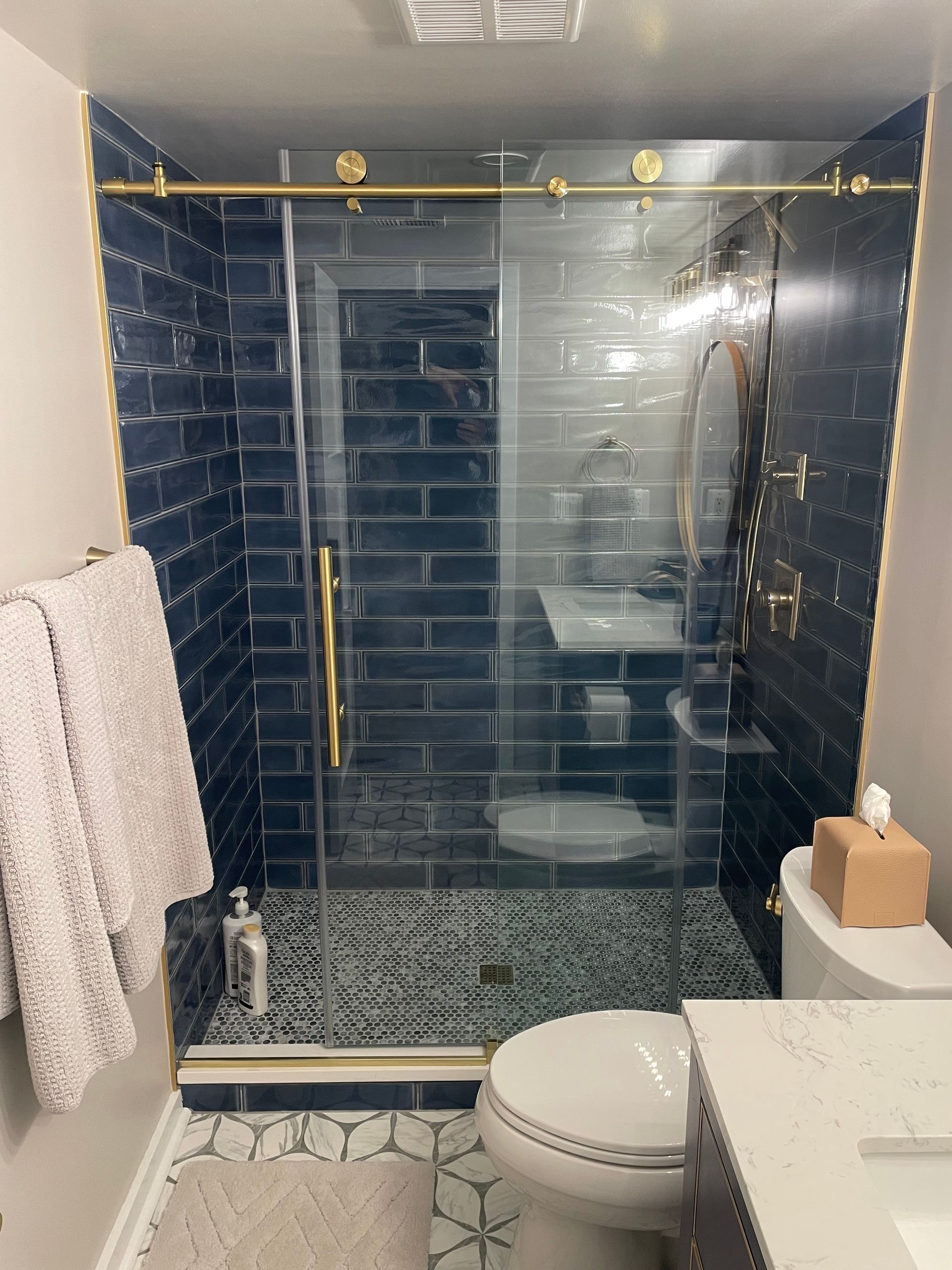 Blue-tiled shower with gold trim and sliding glass doors. A toilet and towels are visible in the bathroom.