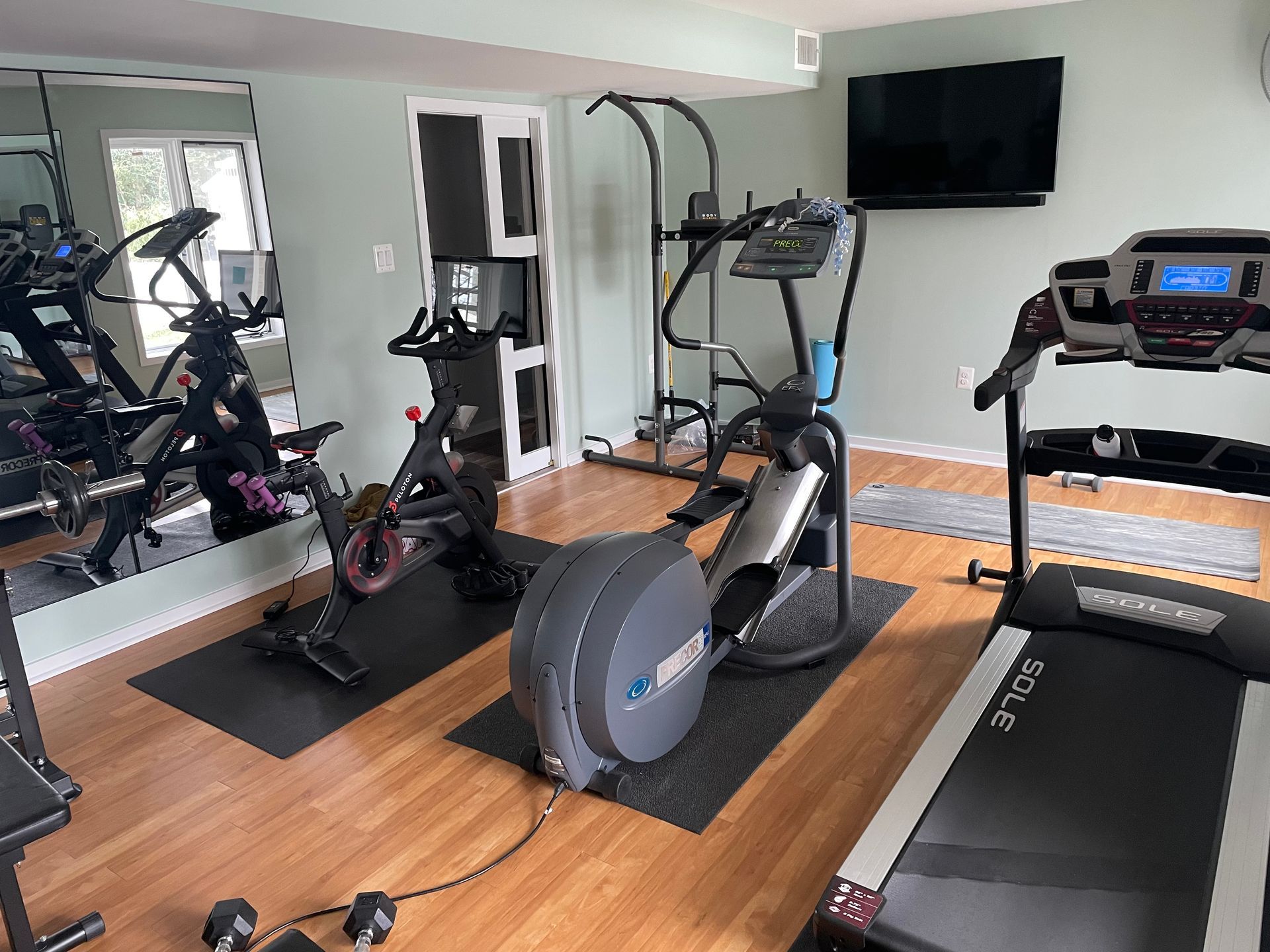 Home gym with treadmill, elliptical, stationary bike, weight bench, mirror, and TV on wall.