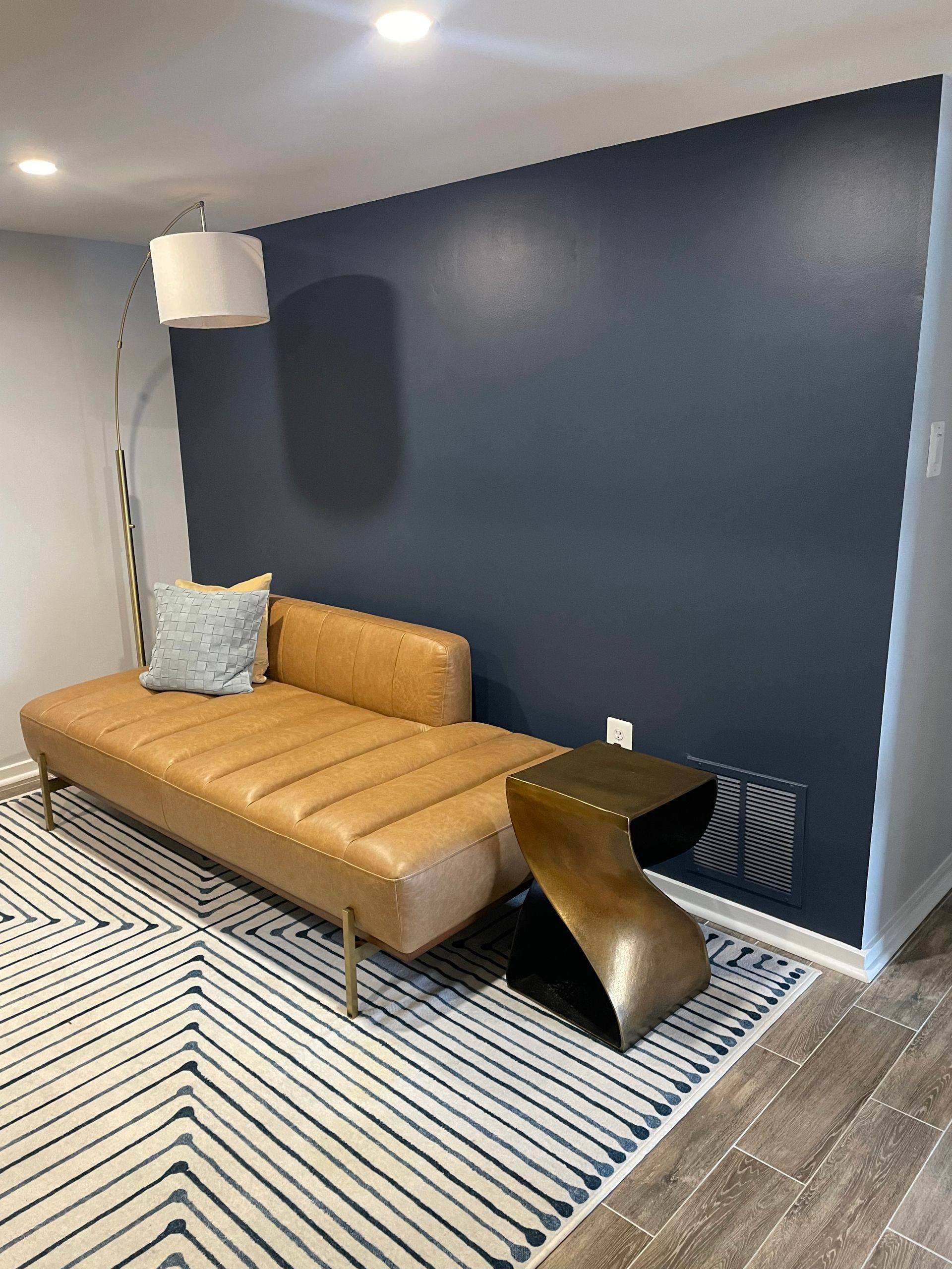 Tan chaise lounge with a throw pillow, side table, and floor lamp against a navy blue wall.