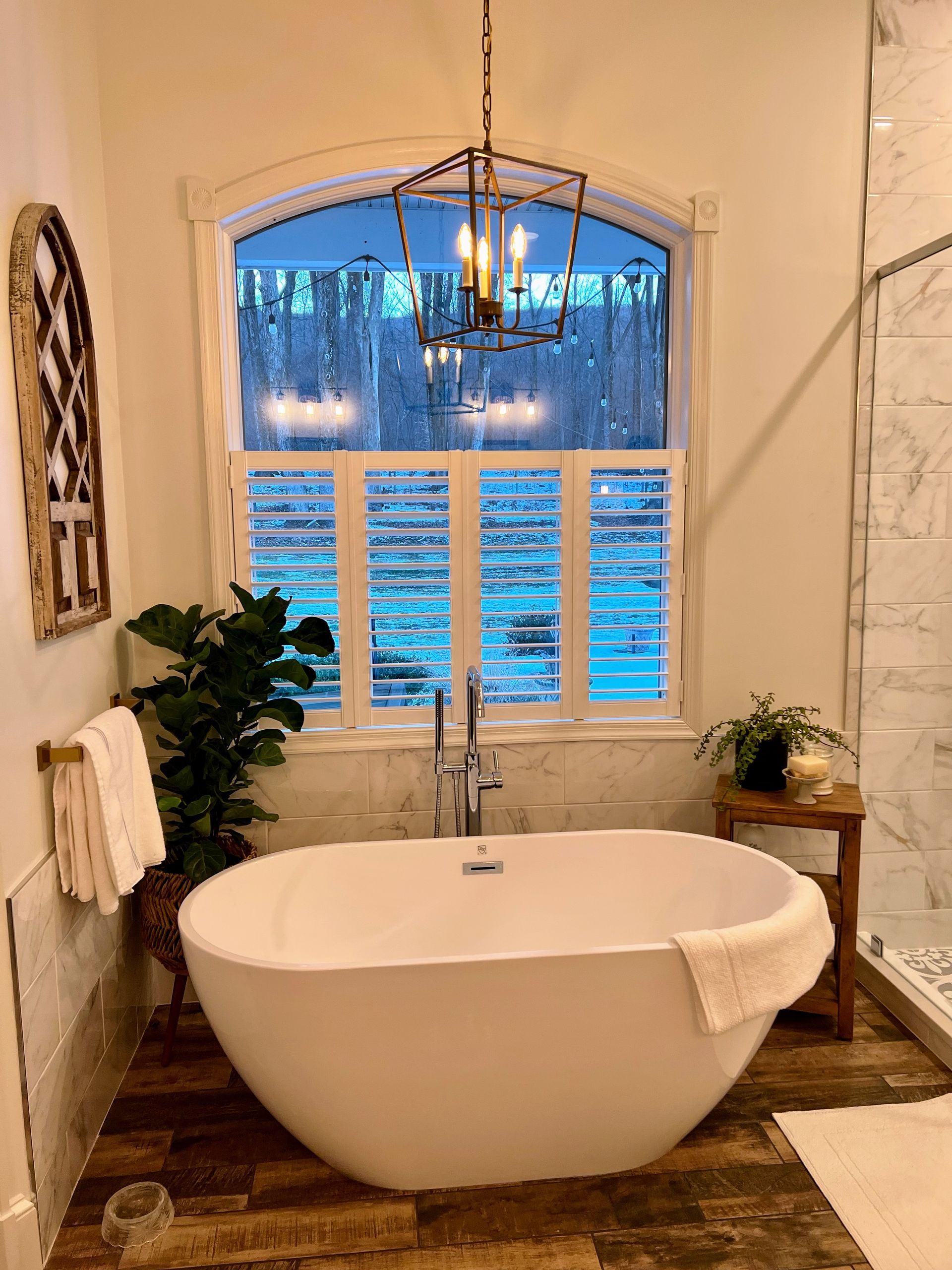 Bathroom with a white freestanding tub, large window with shutters, and a decorative chandelier.