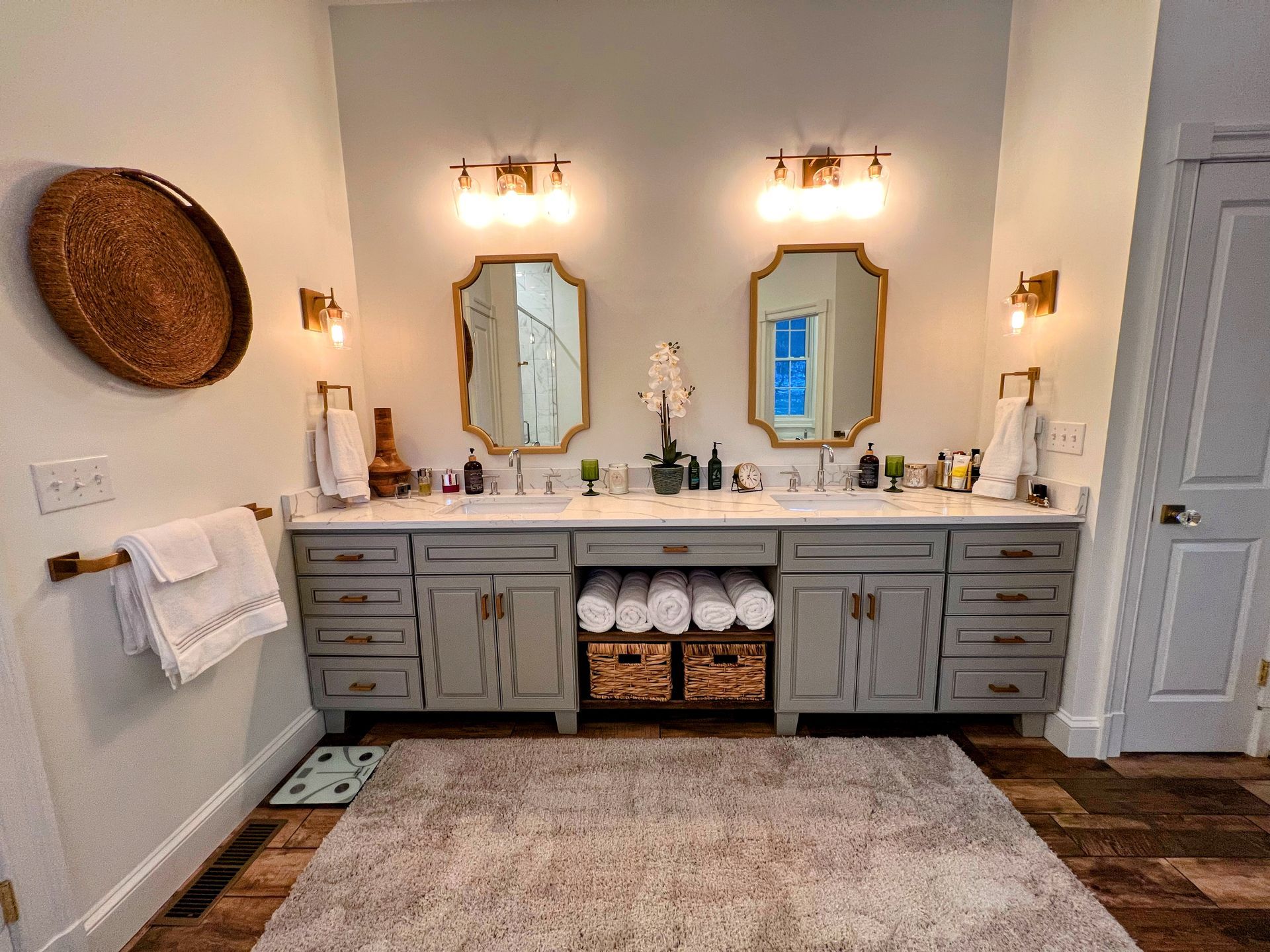 Bathroom with gray vanity, two gold-framed mirrors, and a woven rug.