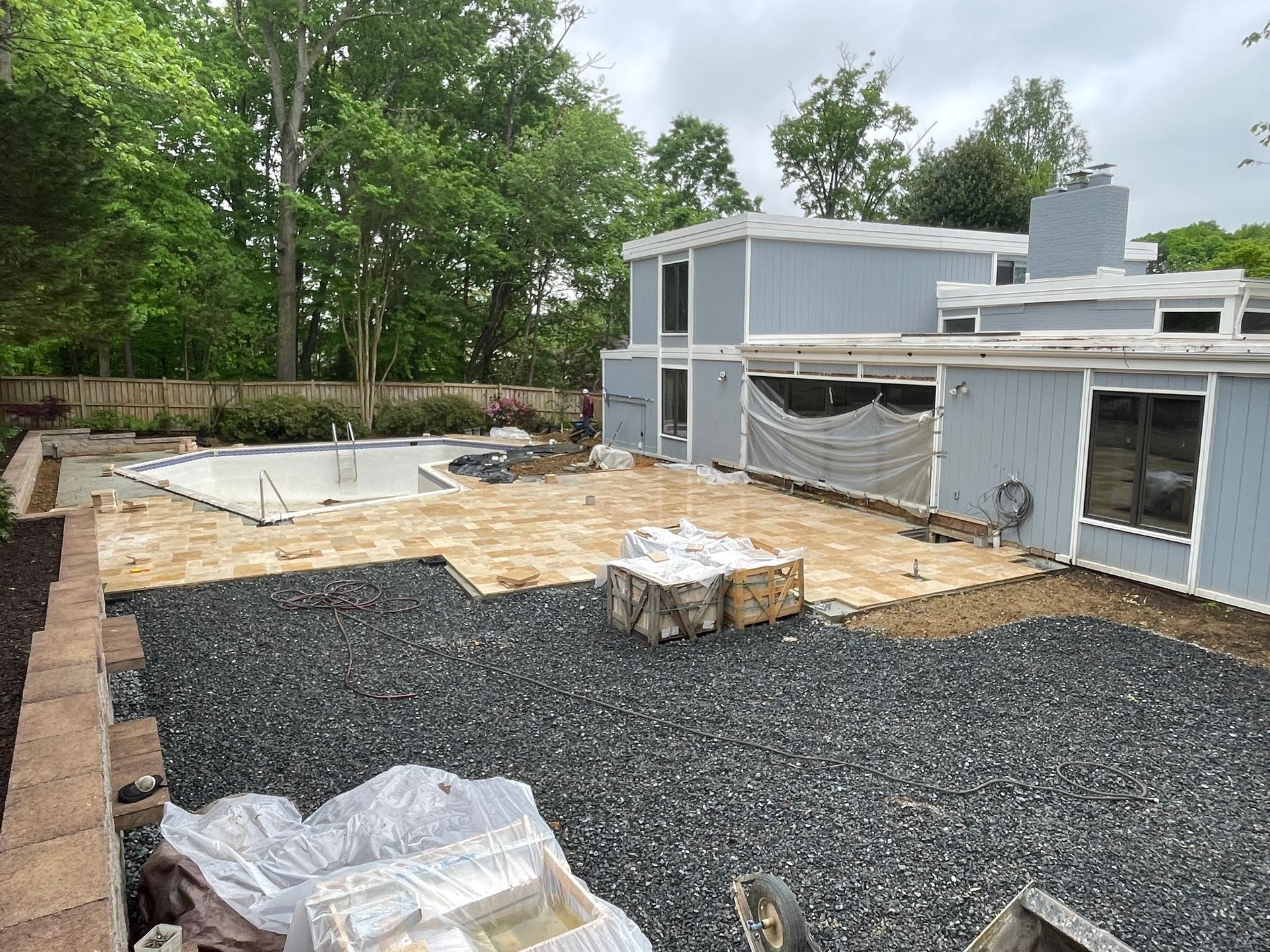 Backyard with pool, construction in progress; gravel, pavers, house painted blue, trees in background.