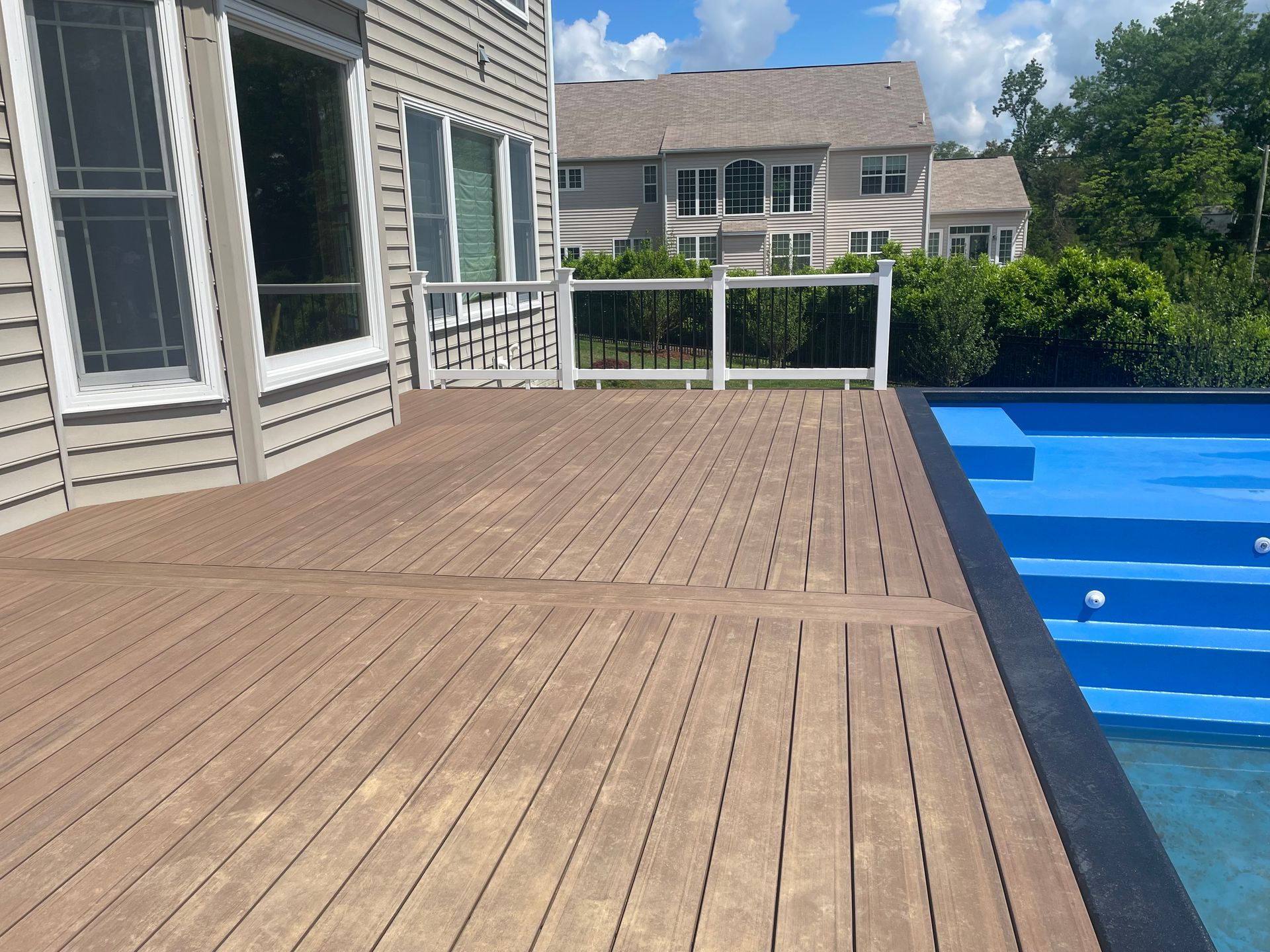 Wooden deck next to a blue pool, a white railing, and a house on a sunny day.