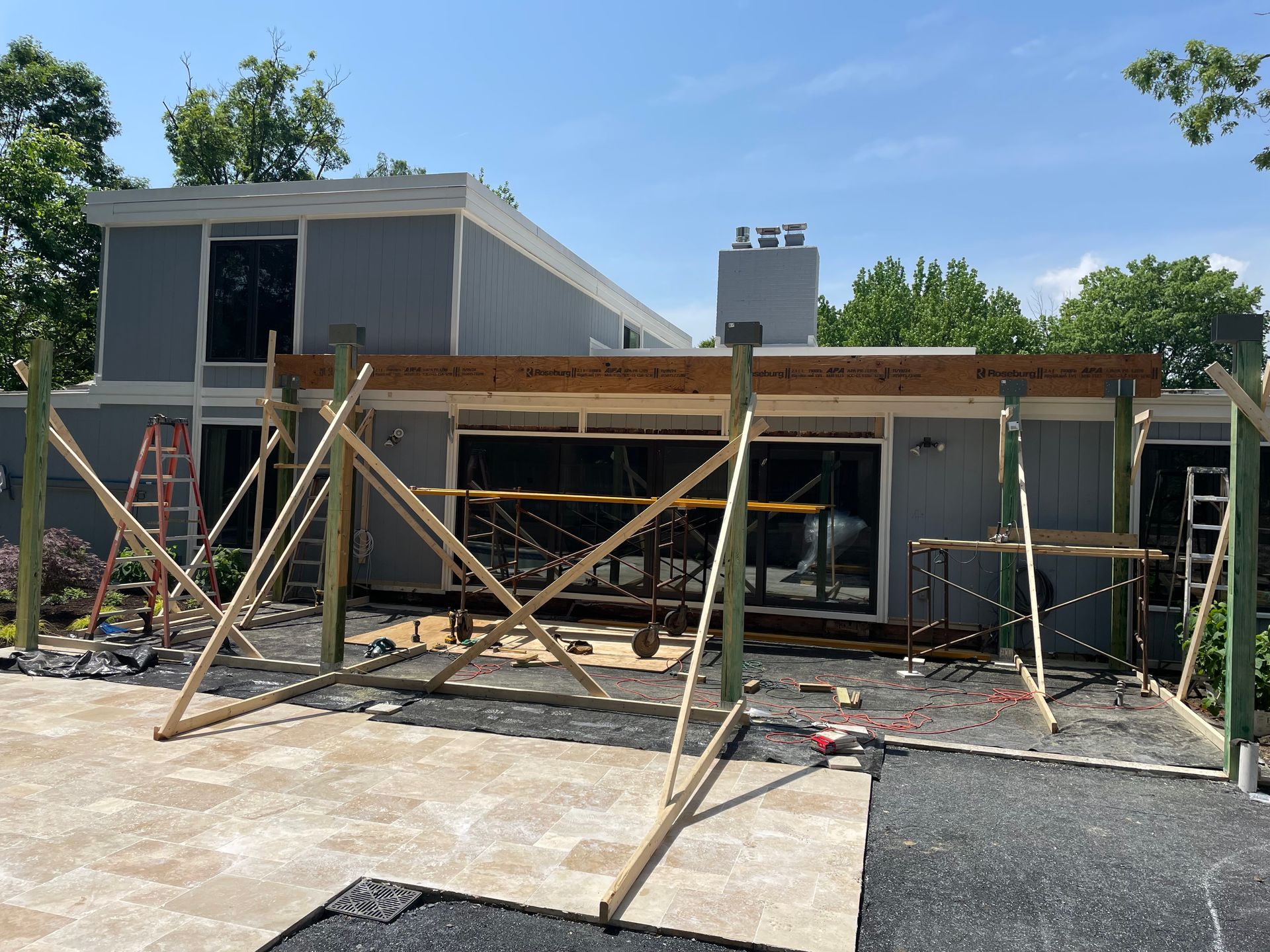 Construction of a wooden pergola attached to a gray house, with supporting scaffolding on a paved patio.