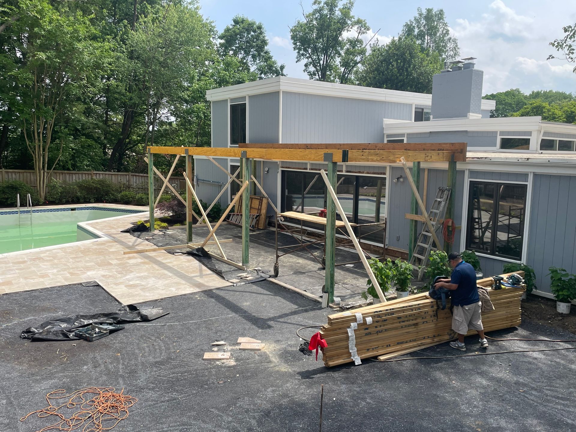 A house under construction. A person is working on a wooden patio roof. Pool and trees in the background.