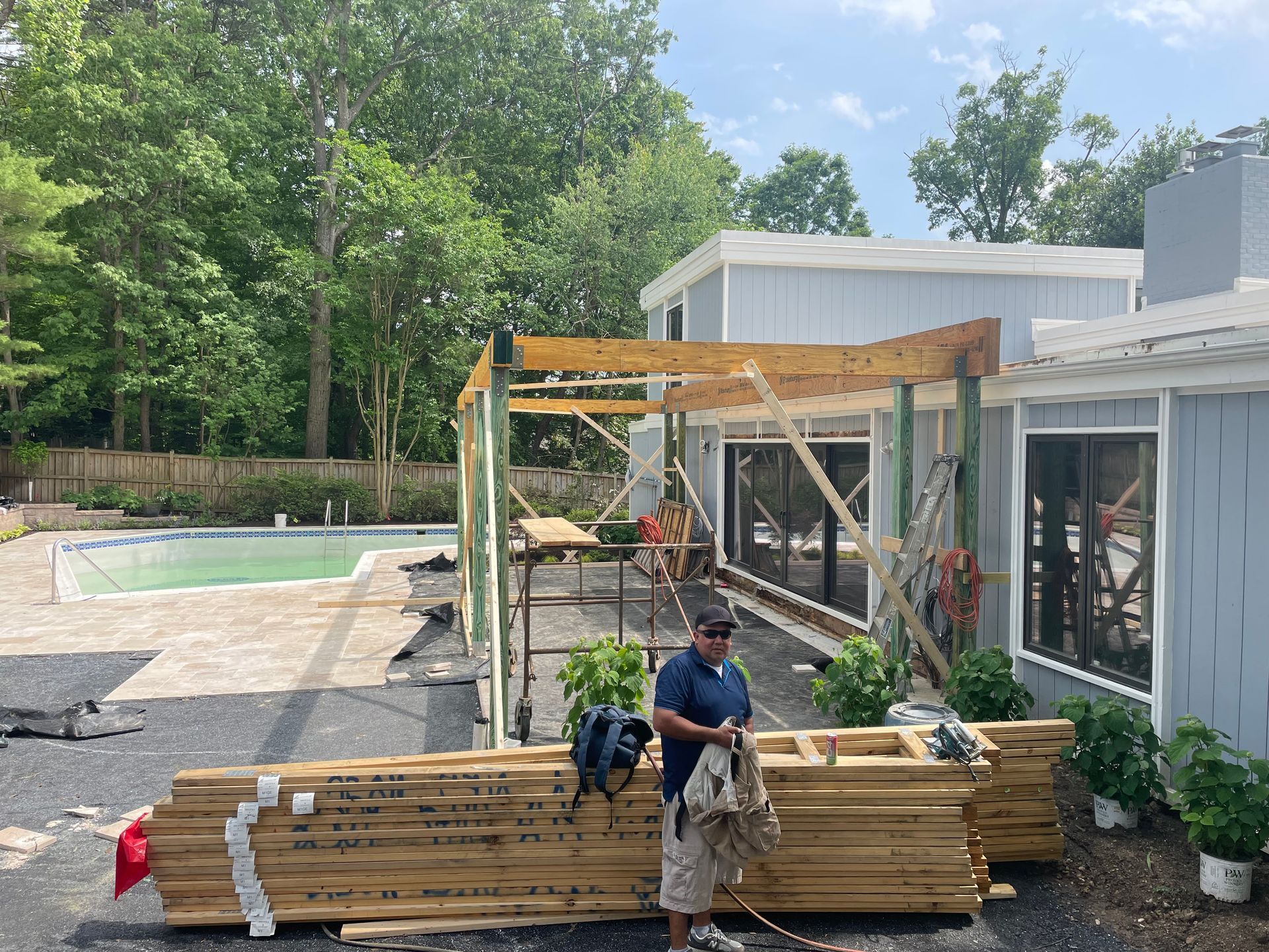 Construction of a patio cover on a light blue house near a pool. A worker stands near lumber.