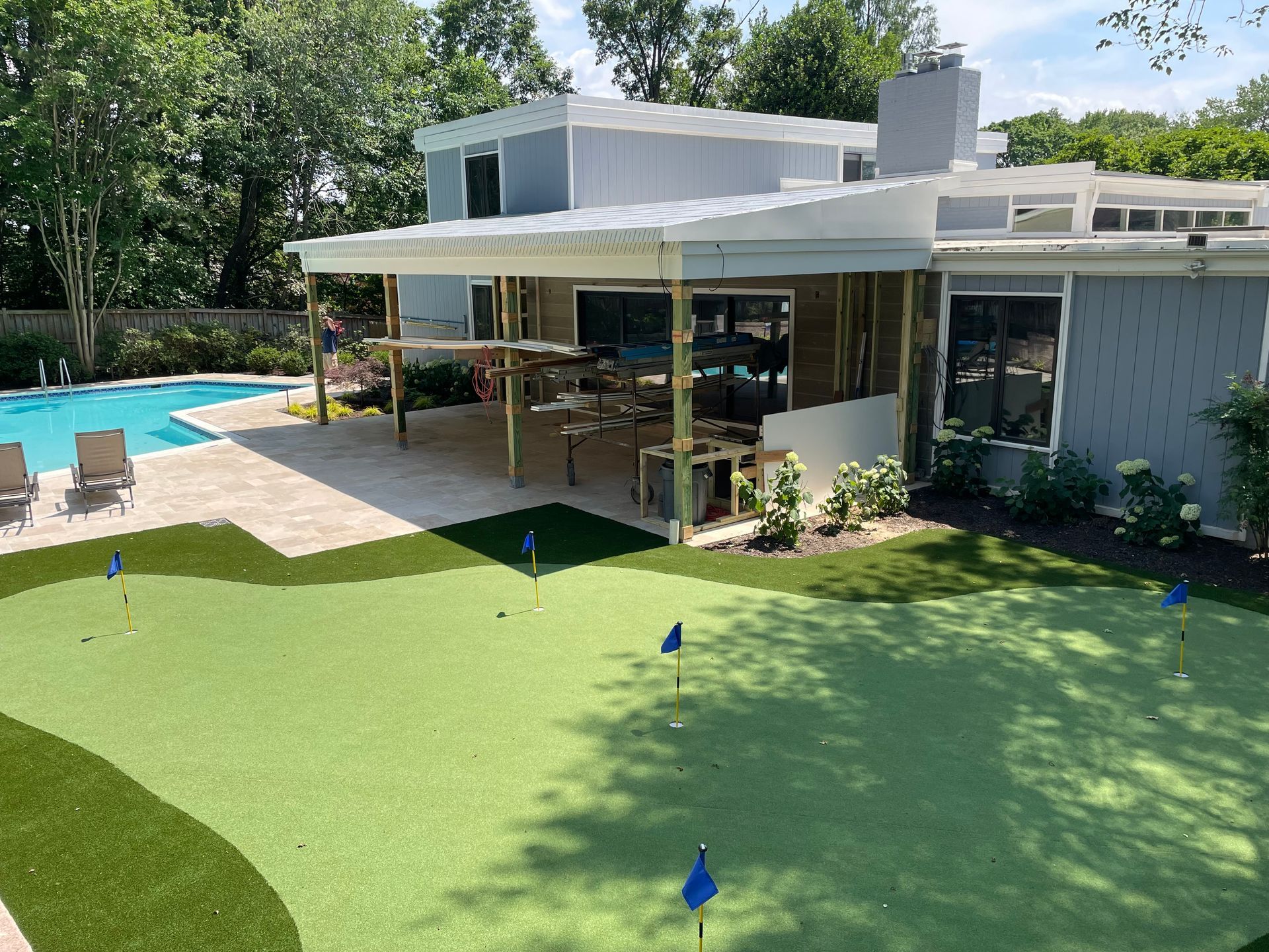 Backyard with a pool, putting green, patio, and modern house. Green grass, blue flags, sunny day.