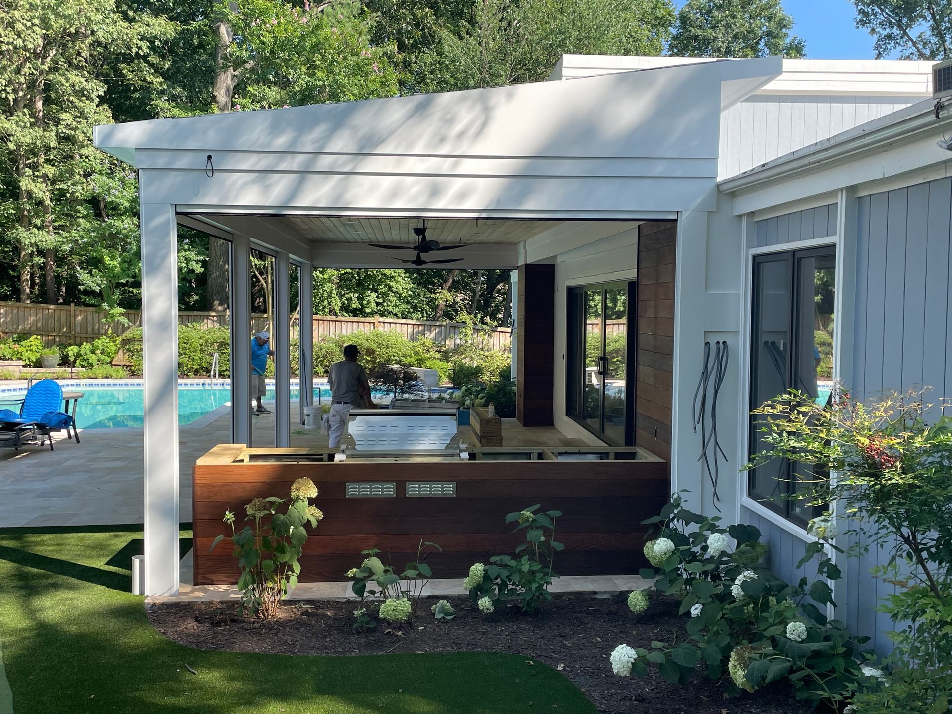 Outdoor kitchen with a grill, overlooking a pool. White pergola, wood accents, and greenery.