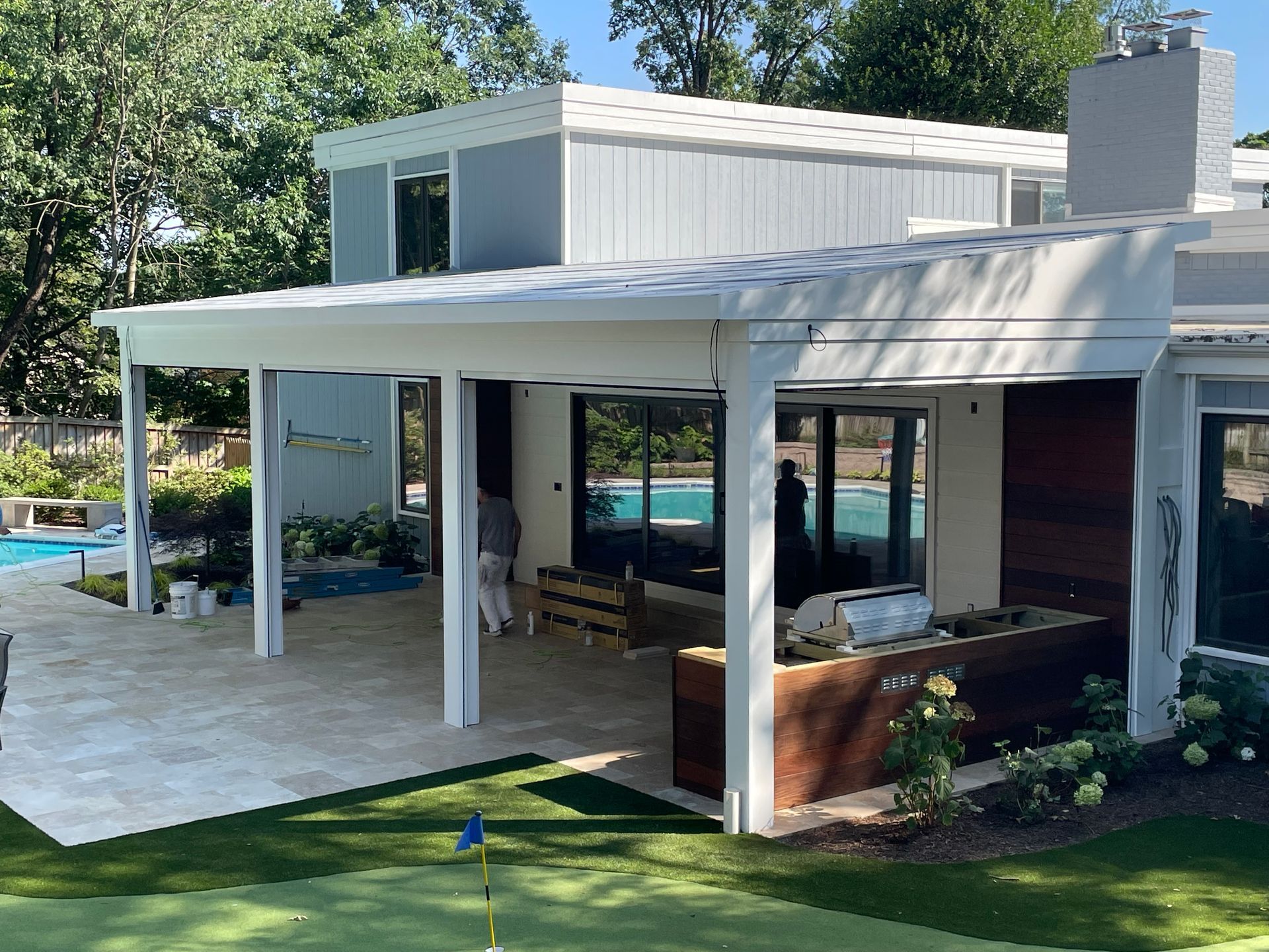White-framed patio with grilling area adjoins a light gray modern house. A swimming pool is visible.
