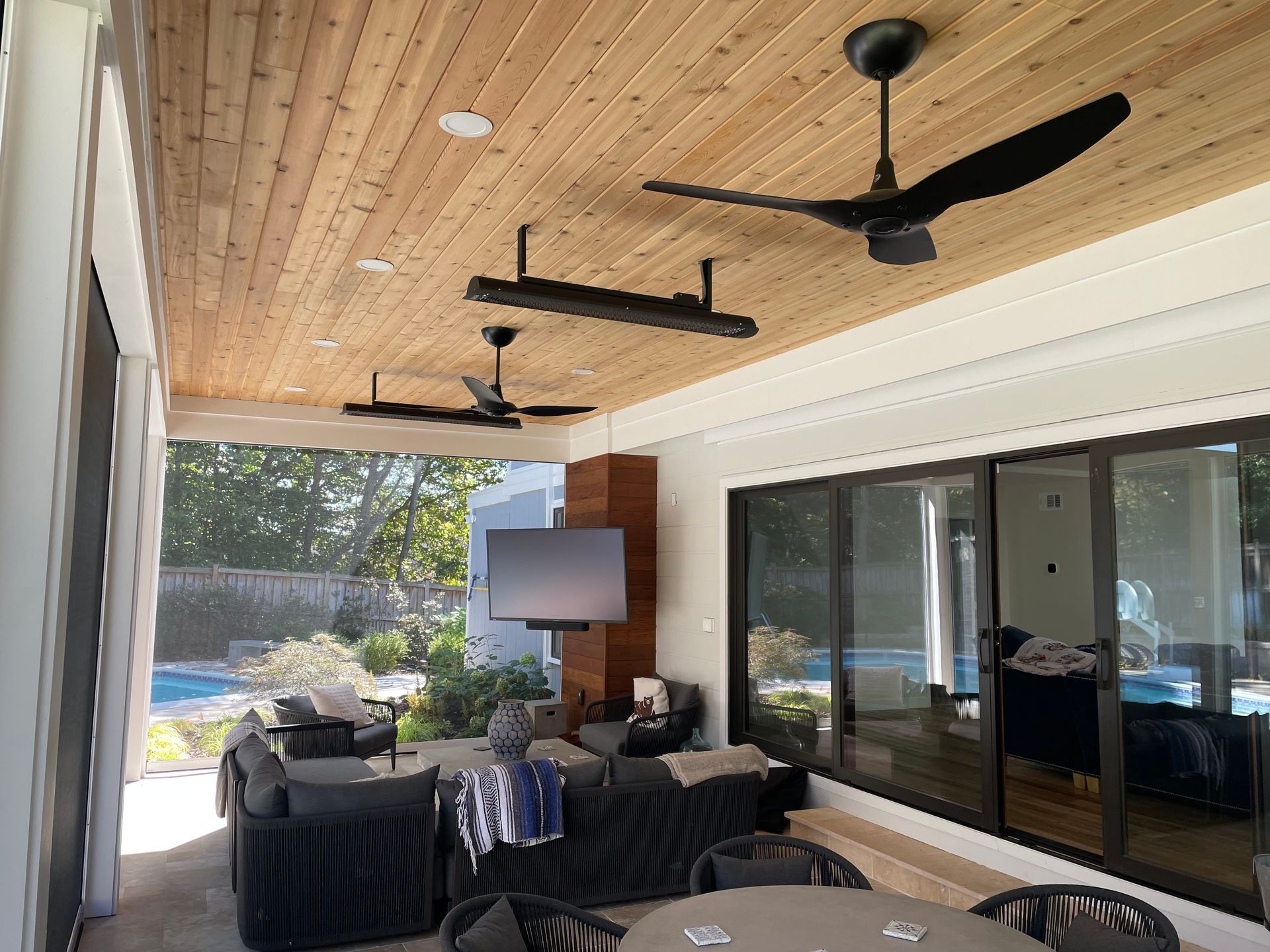 Outdoor patio with wooden ceiling, black ceiling fans, seating area, and sliding glass doors overlooking a pool.