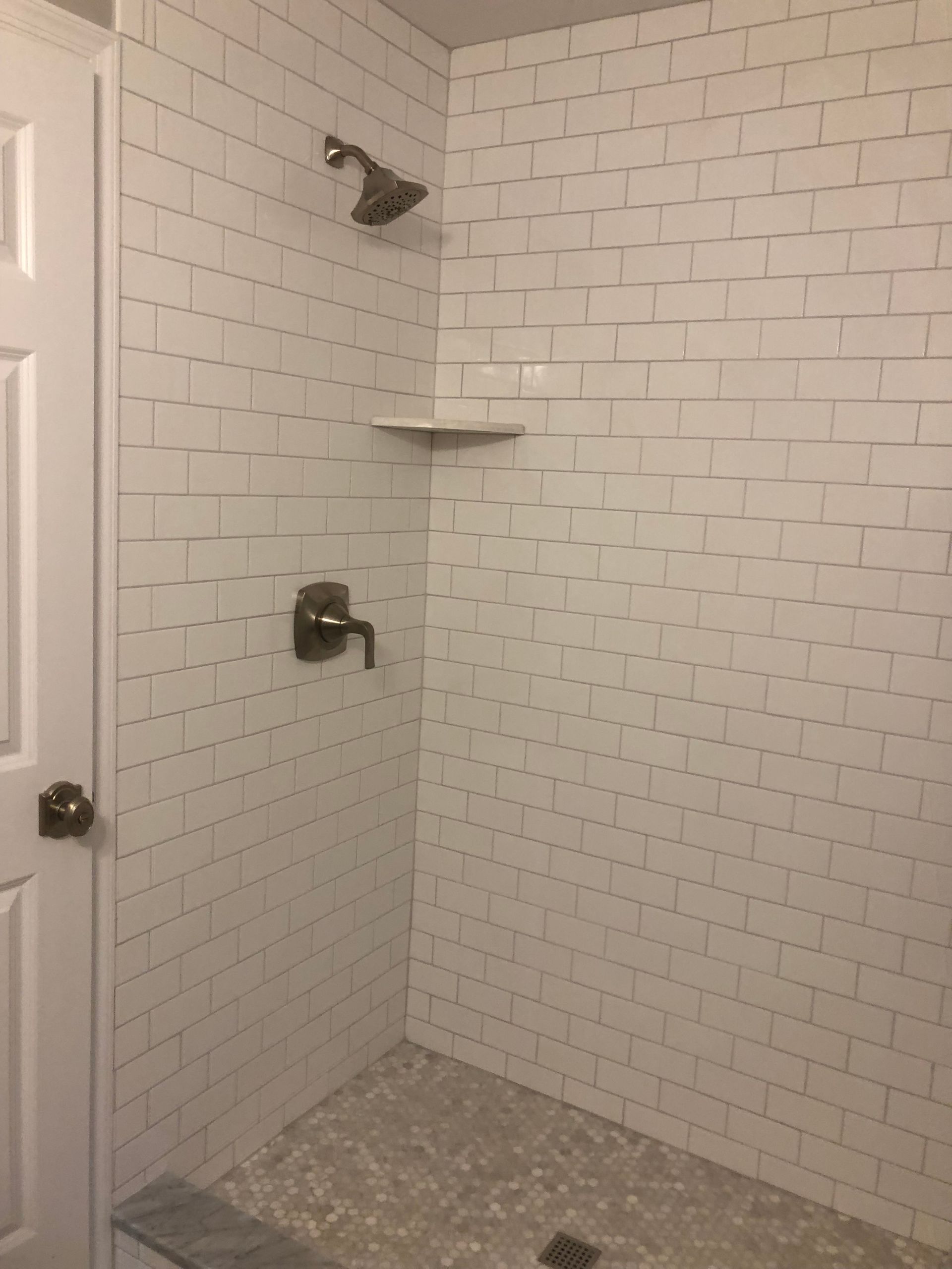 White-tiled shower with a grey pebble floor, a showerhead, a faucet, and a small shelf.