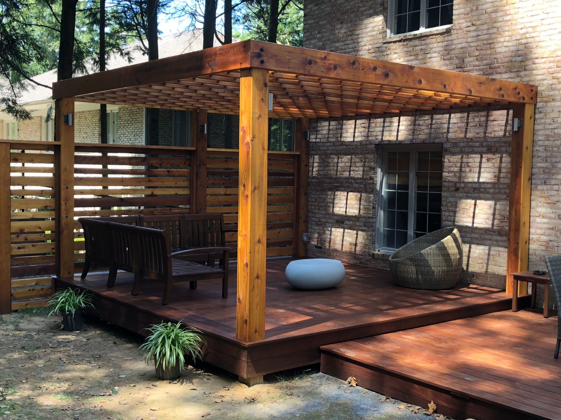 Wooden deck with pergola, seating area, and partial privacy fence next to a brick building.