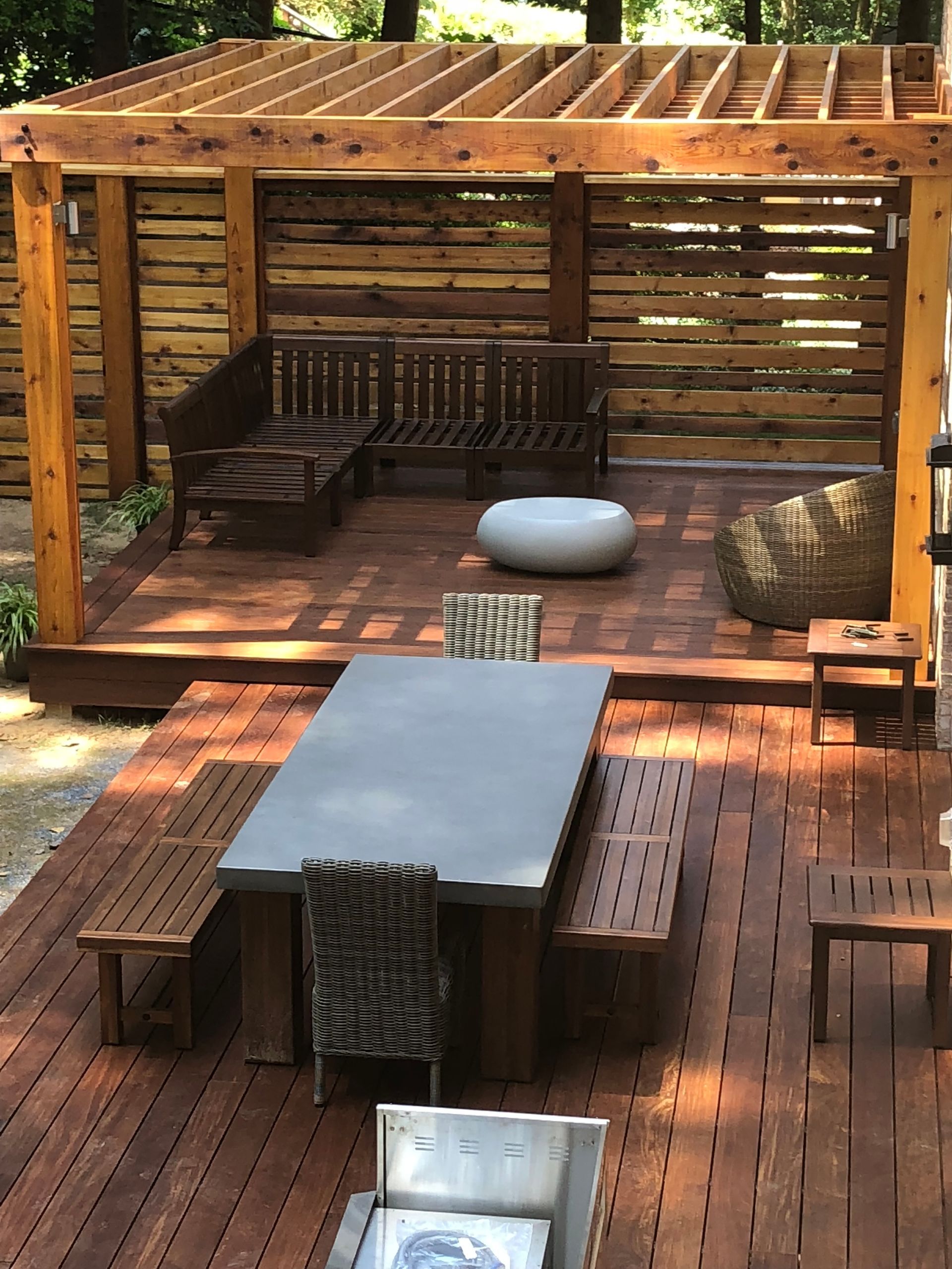 Wooden deck with a pergola, seating areas, and a large gray table.