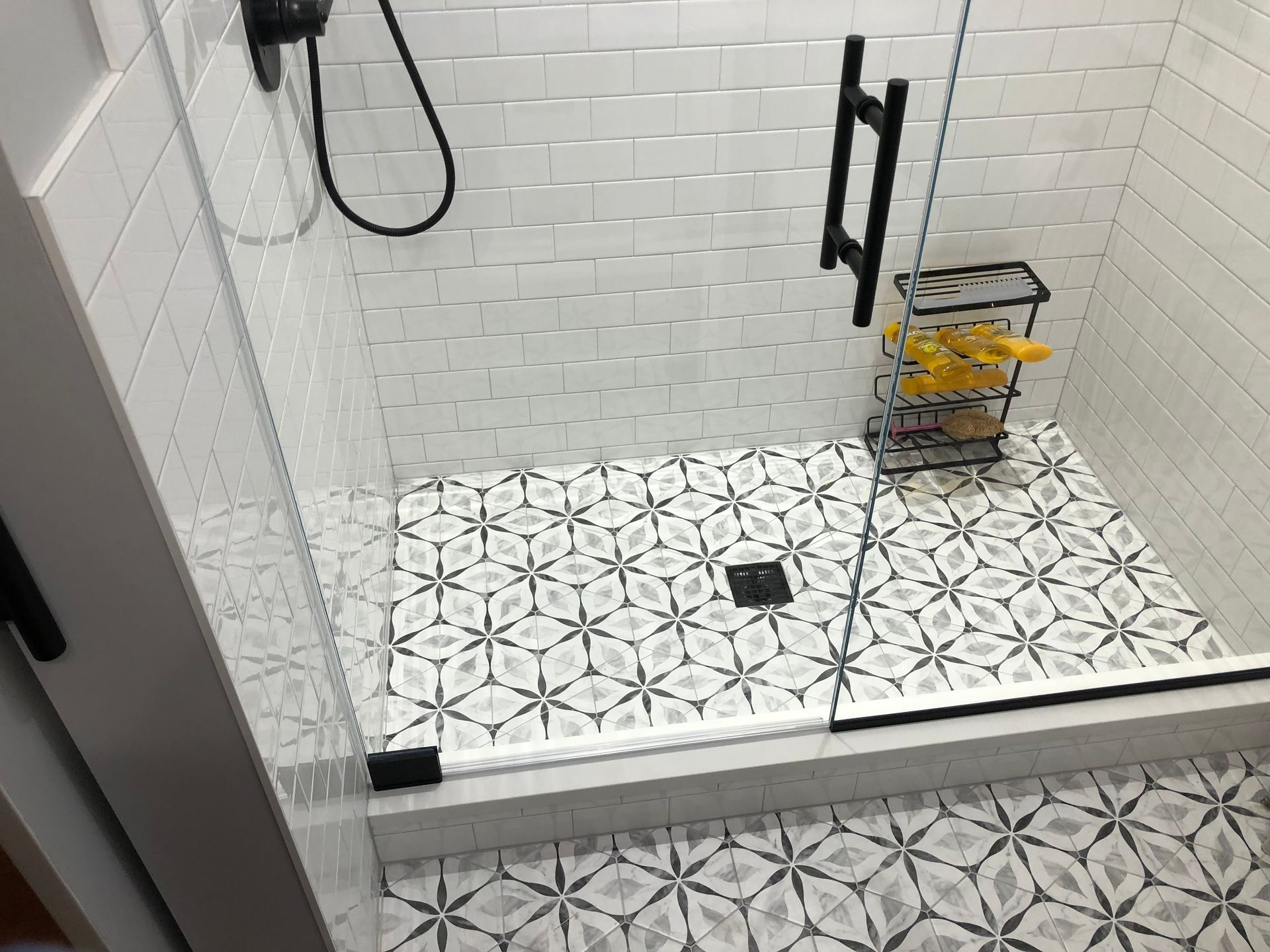 Shower stall with patterned floor and white subway tile walls. Black fixtures, glass door, and caddy.