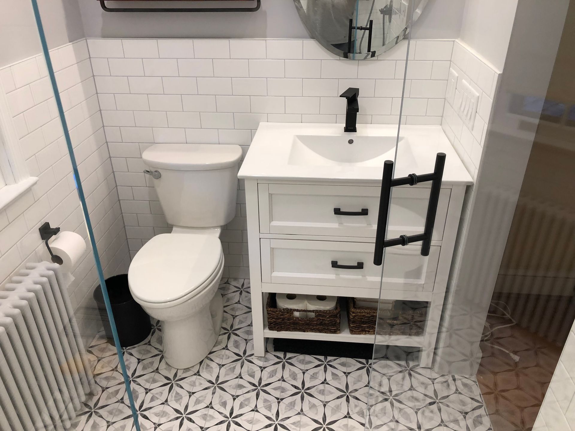 Bathroom with white brick tile, patterned floor, toilet, vanity with black faucet, and shower glass.