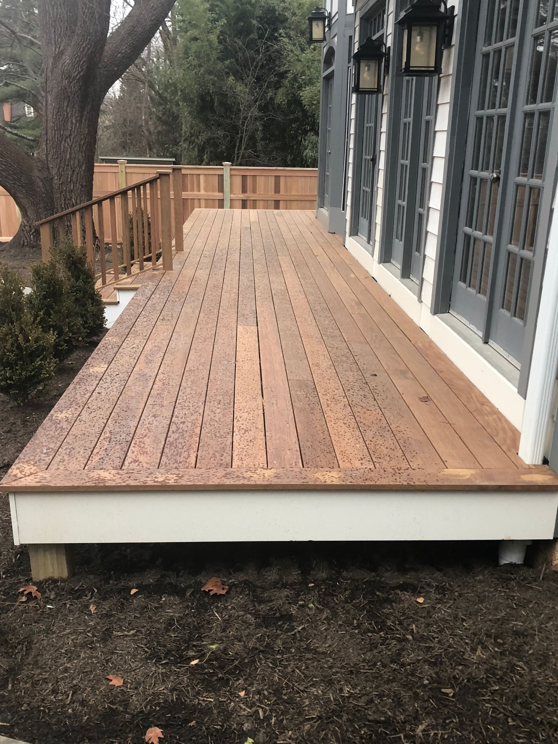 Wooden deck extends from a building. Dark wood planks with spacing. Brown soil and shrubs surround it.
