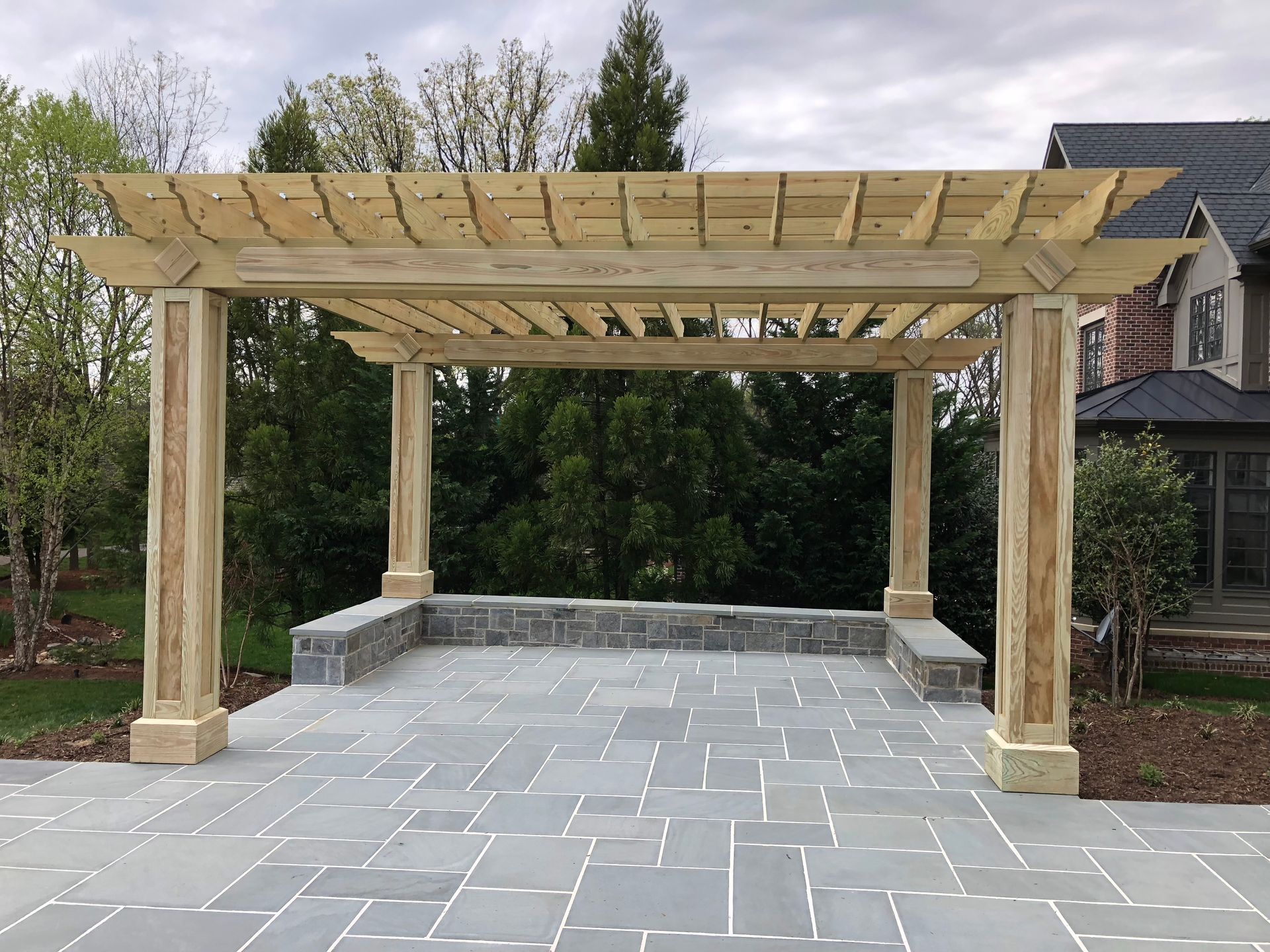 Wooden pergola over a stone patio. Light wood construction with stone accents. Cloudy sky.
